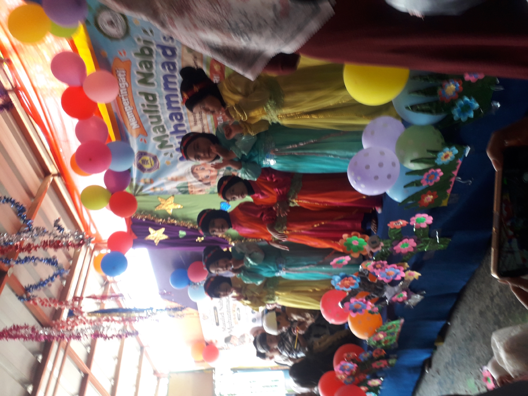 The image captures a festive indoor event, likely a school or community gathering, at an undisclosed location in Cankuang, Indonesia. The scene is decorated with colorful balloons, streamers, and paper flowers, creating a vibrant atmosphere. A banner in the background reads "Memperingati Maulid Nabi Muhammad," indicating the celebration of the Prophet Muhammad's birthday.
In the foreground, a group of children, predominantly girls, are dressed in traditional Islamic attire, including headscarves and long, flowing garments in shades of pink, blue, and yellow. They appear to be either performing on a stage or posing for a photograph. Some are clapping, while others stand with hands clasped.
Behind the performers, an audience of children and adults can be seen, some wearing white skullcaps. The space itself seems to be a covered area, possibly an open-air pavilion or a community hall, with a corrugated metal roof visible. The lighting suggests it is daytime, with sunlight filtering through. A person's hand holding a smartphone is in the lower right corner, actively capturing the moment. The overall impression is one of a lively cultural and religious celebration.