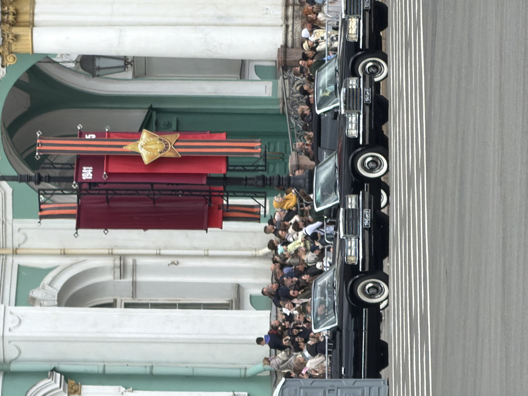 A photograph shows a parade in Saint Petersburg, Russia. There are people watching the parade, as well as several black cars on the right side of the photograph. Decorations are present with "1941" written on a banner.
