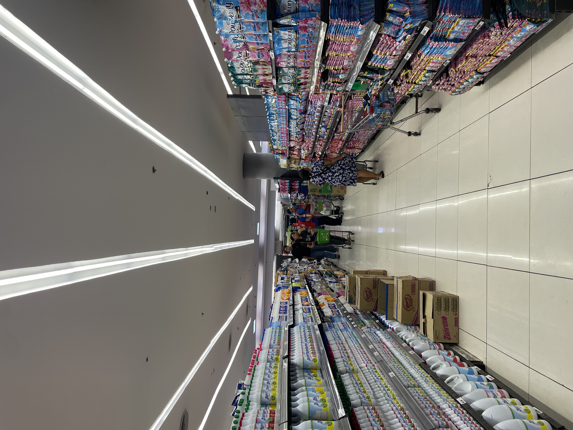 This is a photograph taken inside a brightly lit supermarket aisle in Davao City, Philippines. The aisle is lined with shelves stocked with cleaning products, including bottles of bleach on the left and pouches of fabric conditioner on the right. Several shoppers are visible, some pushing shopping carts and others walking down the aisle. The ceiling features long, linear fluorescent lights. The floor is tiled with large, light-colored tiles. Boxes of cleaning products are stacked on the floor in front of some shelves. The overall atmosphere suggests a typical retail environment for household goods.
