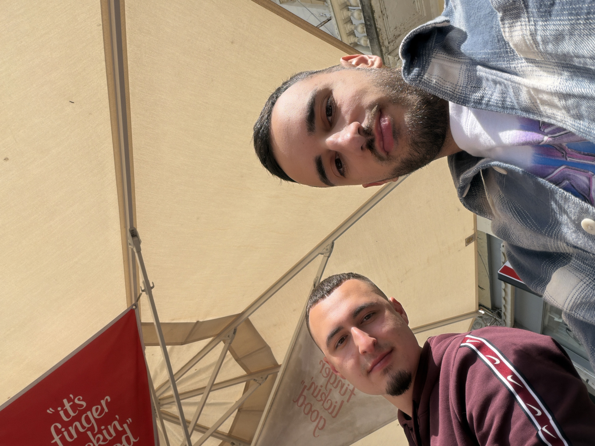 The image captures two men from a low-angle perspective, looking upwards, likely taken as a selfie or by someone positioned below them. They are situated beneath a large, light-colored awning or umbrella structure, typical of an outdoor café or restaurant setting in Plovdiv, Bulgaria.
The man in the upper part of the frame has dark hair and a beard, wearing a blue and white plaid shirt over a white t-shirt with a colorful graphic design. He looks directly into the camera with a neutral expression. The second man, in the lower part of the frame and in sharper focus, has short dark hair and a goatee. He is wearing a maroon hoodie featuring a distinctive white and red stripe with white Nike swoosh logos running along the shoulder and sleeve. He also looks directly at the camera with a slight, subtle smile.
The awning, made of light tan fabric panels, is supported by metal frames, and its structure is clearly visible. Bright sunlight illuminates the scene, casting highlights on the men's faces and the fabric, suggesting a clear, sunny daytime.
Visible text provides cues about the location. On a red banner in the lower-left corner, partial text reads "it's finger" and "ckin'," which strongly implies the slogan "it's finger lickin' good." Further confirming this, a light-colored banner on the mid-right shows the words "good" and "lickin'" displayed upside down and mirrored, consistent with viewing text from below through a translucent material or a specific flag design. These details suggest the establishment is likely associated with KFC or a similar food service. The backdrop hints at an urban environment with building structures just beyond the awning.