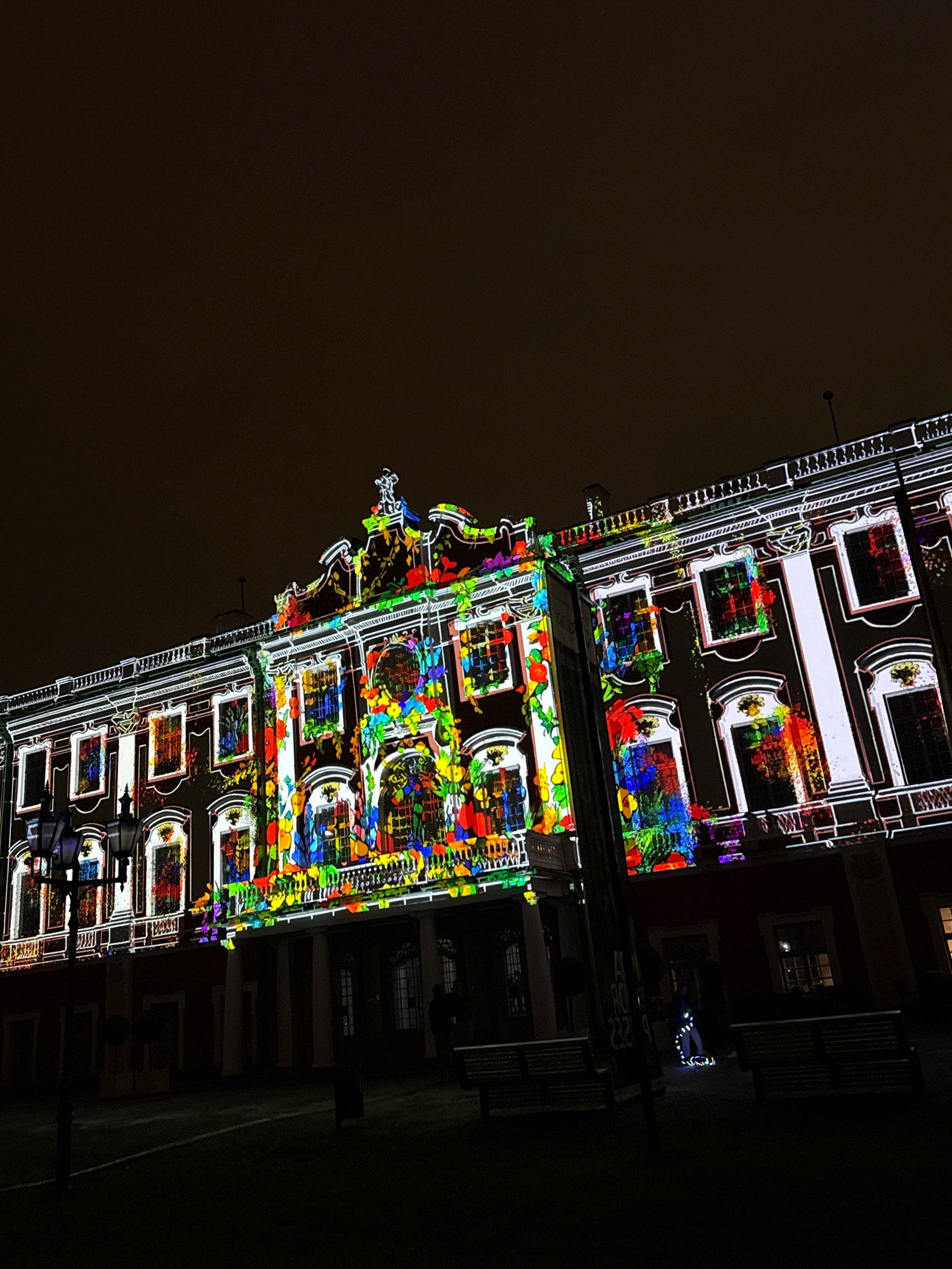 The image shows a building in Tallinn, Estonia, illuminated by a vibrant projection mapping display at night. The building, likely a historical structure, features ornate architecture with multiple windows and decorative elements. The projection displays a colorful and dynamic pattern of abstract shapes, floral designs, and possibly animated figures across its facade, creating a striking visual effect against the dark sky. In the foreground, a lamppost with multiple lights stands on the left side of the image. There are benches in the mid-ground, and a few indistinct figures can be seen in the distance, one of whom appears to be illuminated by a reflective or illuminated element on their clothing. The ground is dark, suggesting a paved area or park surrounding the building. The overall impression is one of a festive or artistic event taking place in the city.