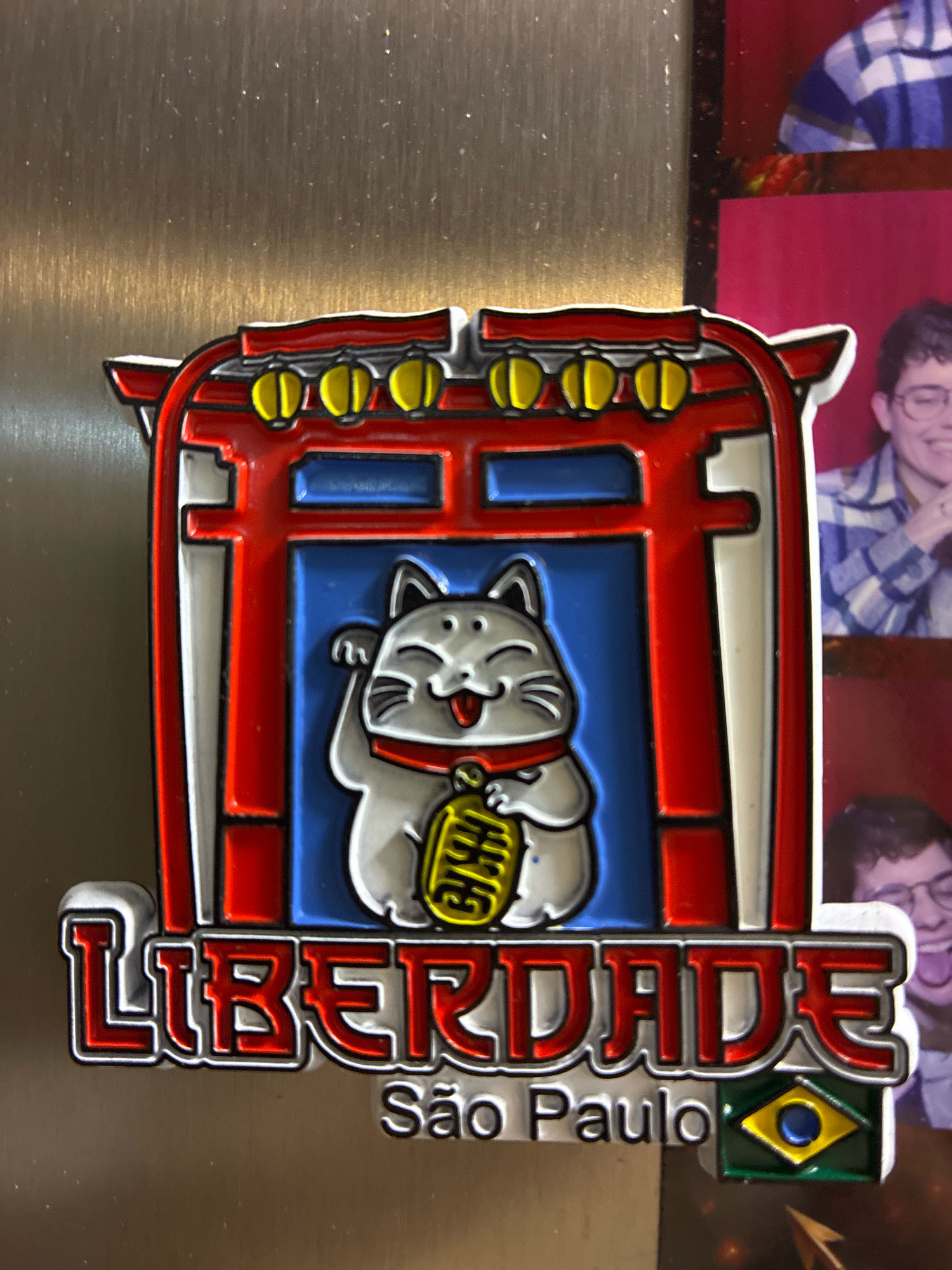A close-up, slightly angled shot shows a colorful magnet on a metallic surface. The magnet depicts a red torii gate with yellow accents, framing a smiling white cat with a red collar and a gold oban coin. Below the cat and gate, the word "LIBERDADE" is emblazoned in red with a white outline, followed by "Sao Paulo" in smaller white text. To the right of "Sao Paulo" is a small Brazilian flag. The background consists of a blurred metallic surface and a portion of what appears to be a photo collage featuring two individuals. The person in the upper portion of the collage is wearing a plaid shirt, and the person in the lower portion is also visible with a blurred background. The overall scene does not provide any information about the city of Curitiba, Brazil.