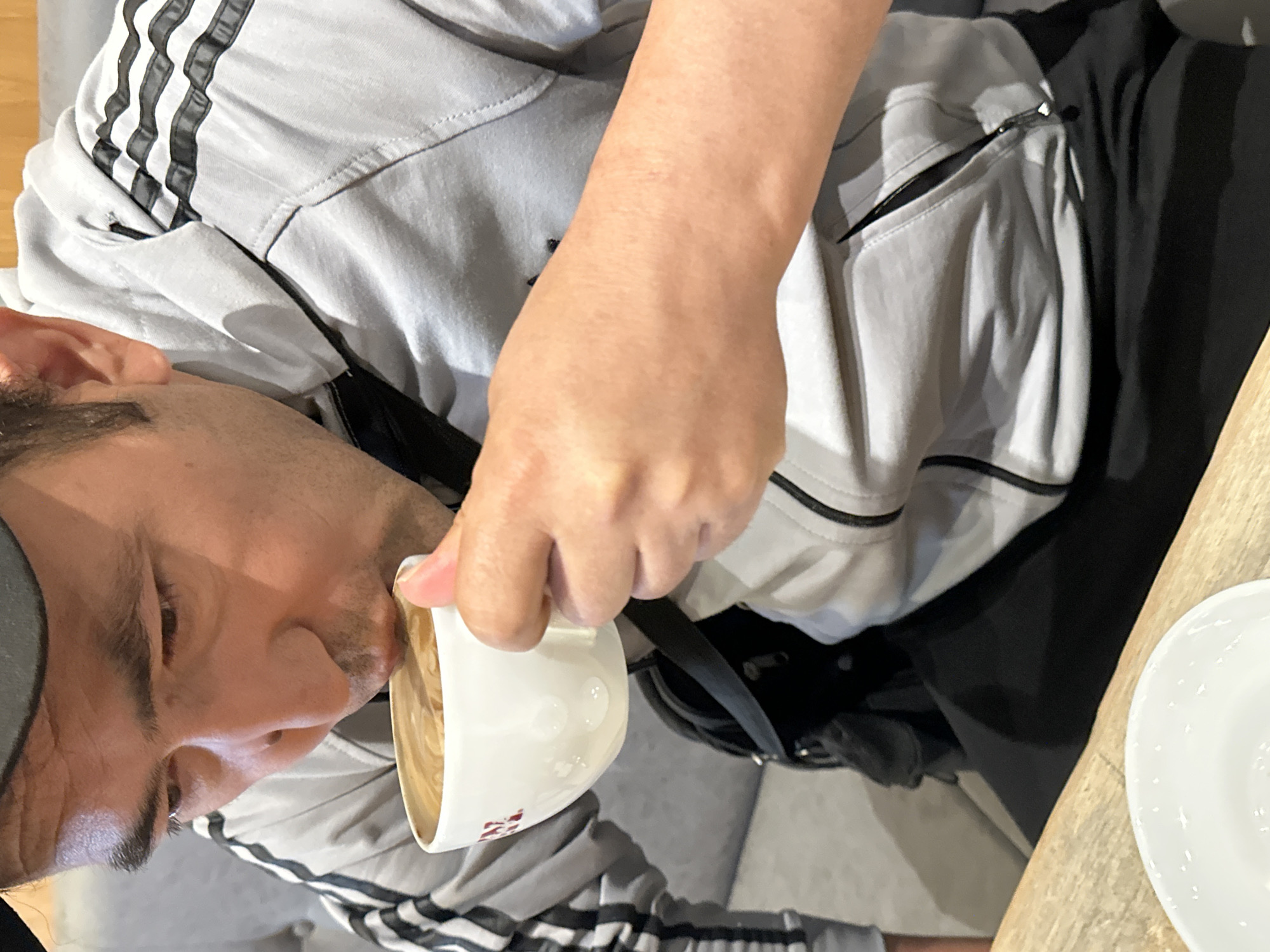A man wearing a grey Adidas jacket with black stripes on the sleeves and a black bandana drinks from a white coffee cup. The coffee has latte art on top. He is sitting on a grey couch in what appears to be a cafe. The image is cropped tightly on the man and his coffee, obscuring most of the environment. The man's face is visible, looking down at the coffee cup as he drinks. The scene appears to be indoors, with a wooden table in the foreground and hints of furniture in the background. The image does not provide any information about a specific location in Krefeld, Germany.