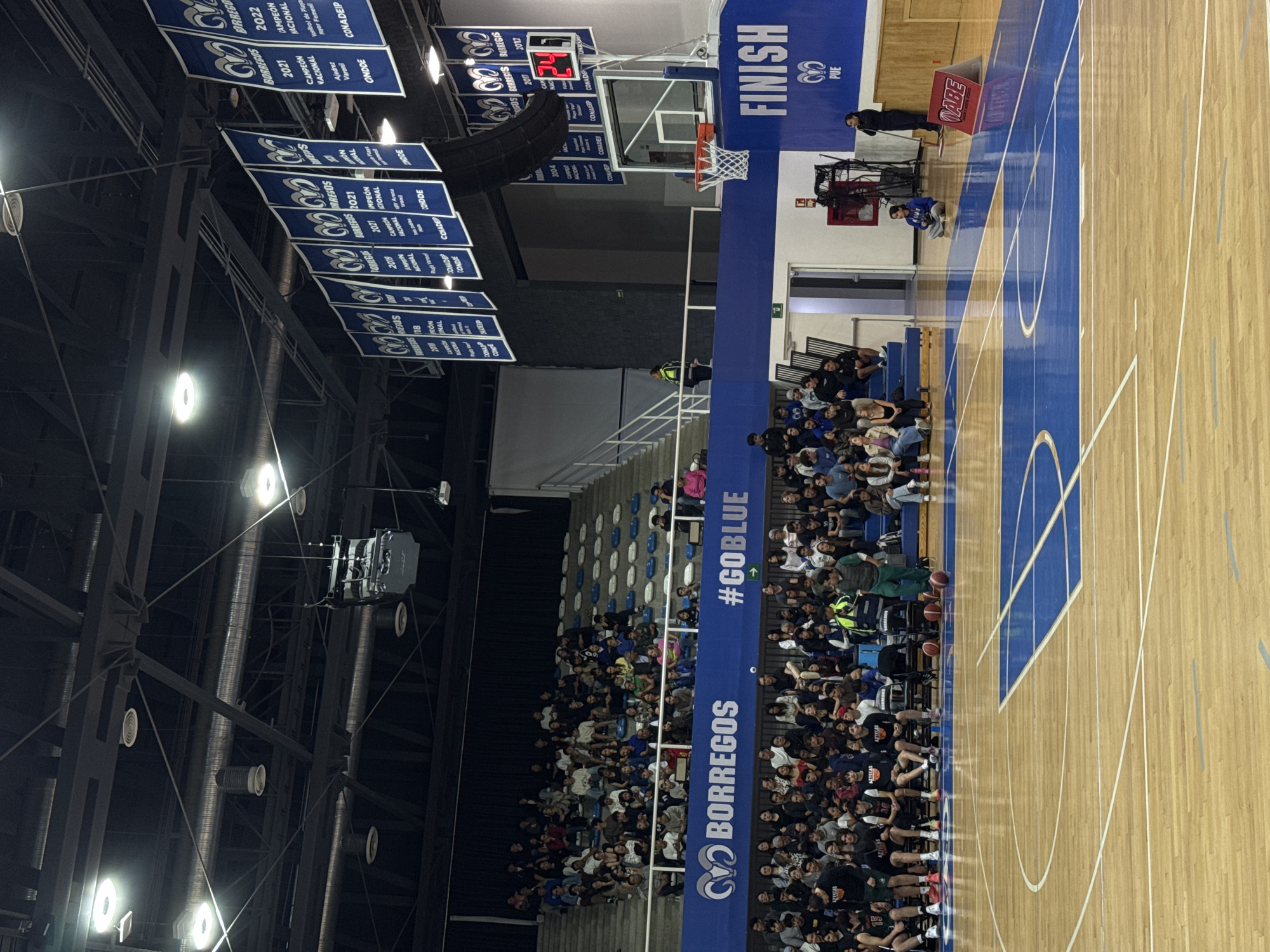The image is an angled, indoor shot of a basketball game with spectators in bleacher seating. Banners hang from the ceiling, displaying sports achievements. The basketball court has blue markings and text on the court divider with "#GOBLUE." A scoreboard indicates the game is in progress. The audience fills the lower level of the seating. A basketball hoop is visible at one end of the court. The environment is an indoor sports arena. Text on the banner states the names of the teams, “Borregos,” and “Aztecas.” Text on the banner states “Heroica Puebla de Zaragoza, Mexico.”