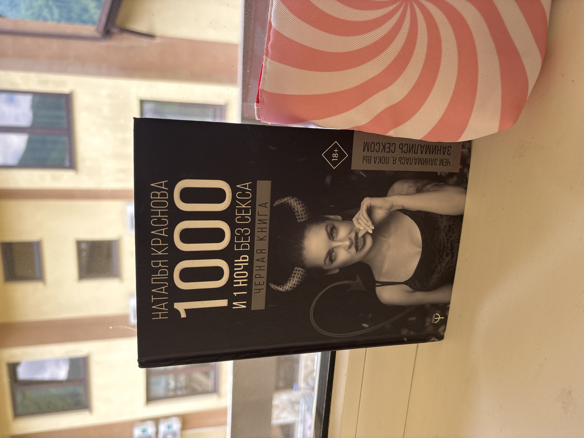 A book titled "1000 and 1 Night Without Sex" by Natalia Krasnova is displayed on a windowsill in Krasnaya Polyana, Russia. The book cover features an image of a woman with devil horns and the "18+" symbol. To the right of the book is a pink and white striped object, possibly a bag or pouch. The background shows windows of a building with a mountainous landscape visible through them.
