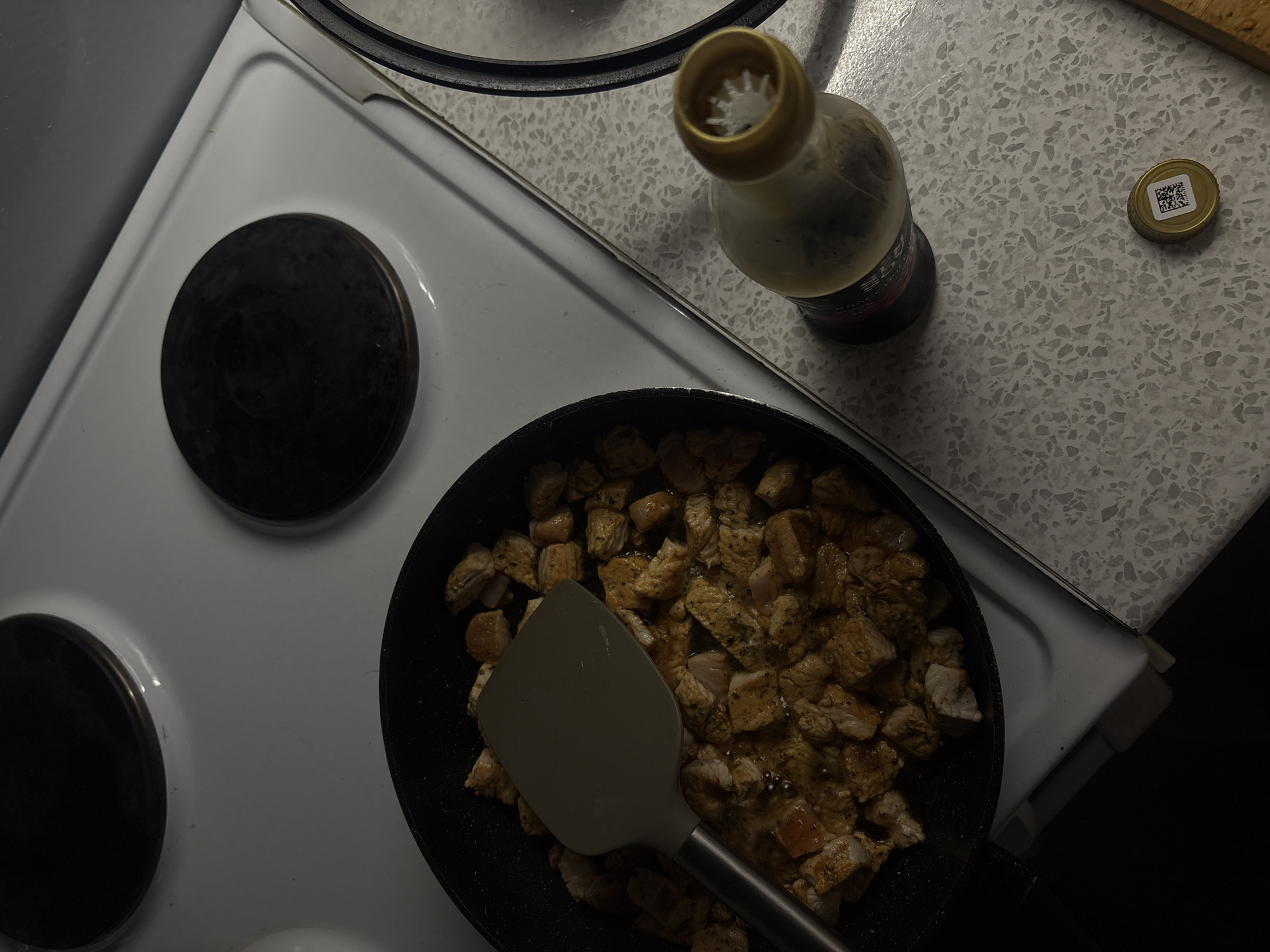 In an indoor residential kitchen, likely in Khimki, Russia, a black frying pan containing diced, cooked chicken is centered on a white electric stovetop. A grey spatula is placed inside the pan, resting on the chicken. To the right of the stovetop, on a speckled countertop, sits an open bottle of dark liquid with a gold cap, which is lying nearby and also features a QR code. The ambient lighting suggests it is either evening or the room is dimly lit. The background shows portions of the stovetop burners and the upper edge of the countertop.