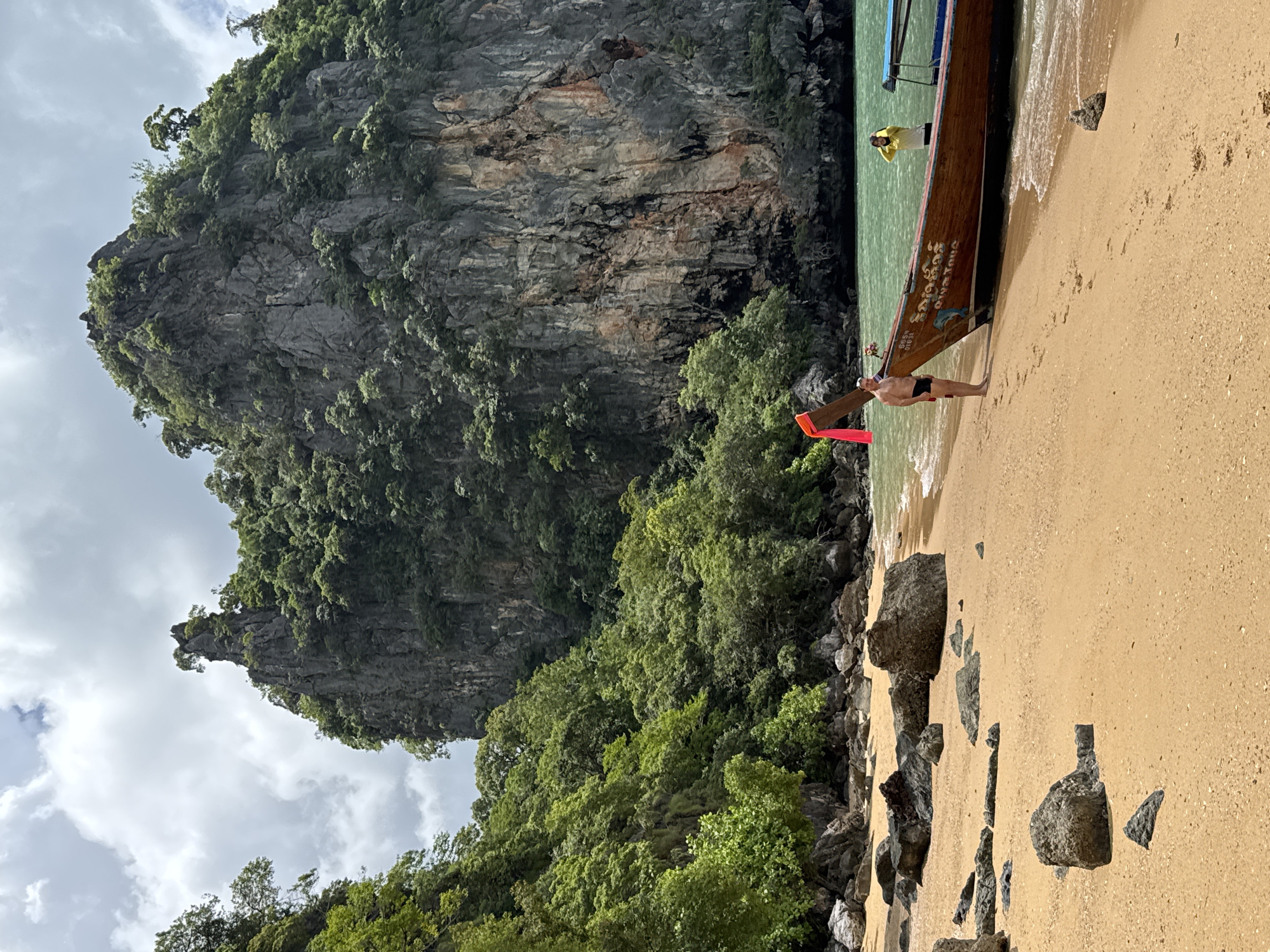 A man and a woman are standing on a sandy beach in Khlong Khian, Thailand. The man is wearing black swim trunks and is standing next to a wooden boat. The woman is wearing a yellow shirt and is standing in the water. In the background, there is a large rock formation covered in trees. The water is a clear turquoise color. The sky is cloudy.