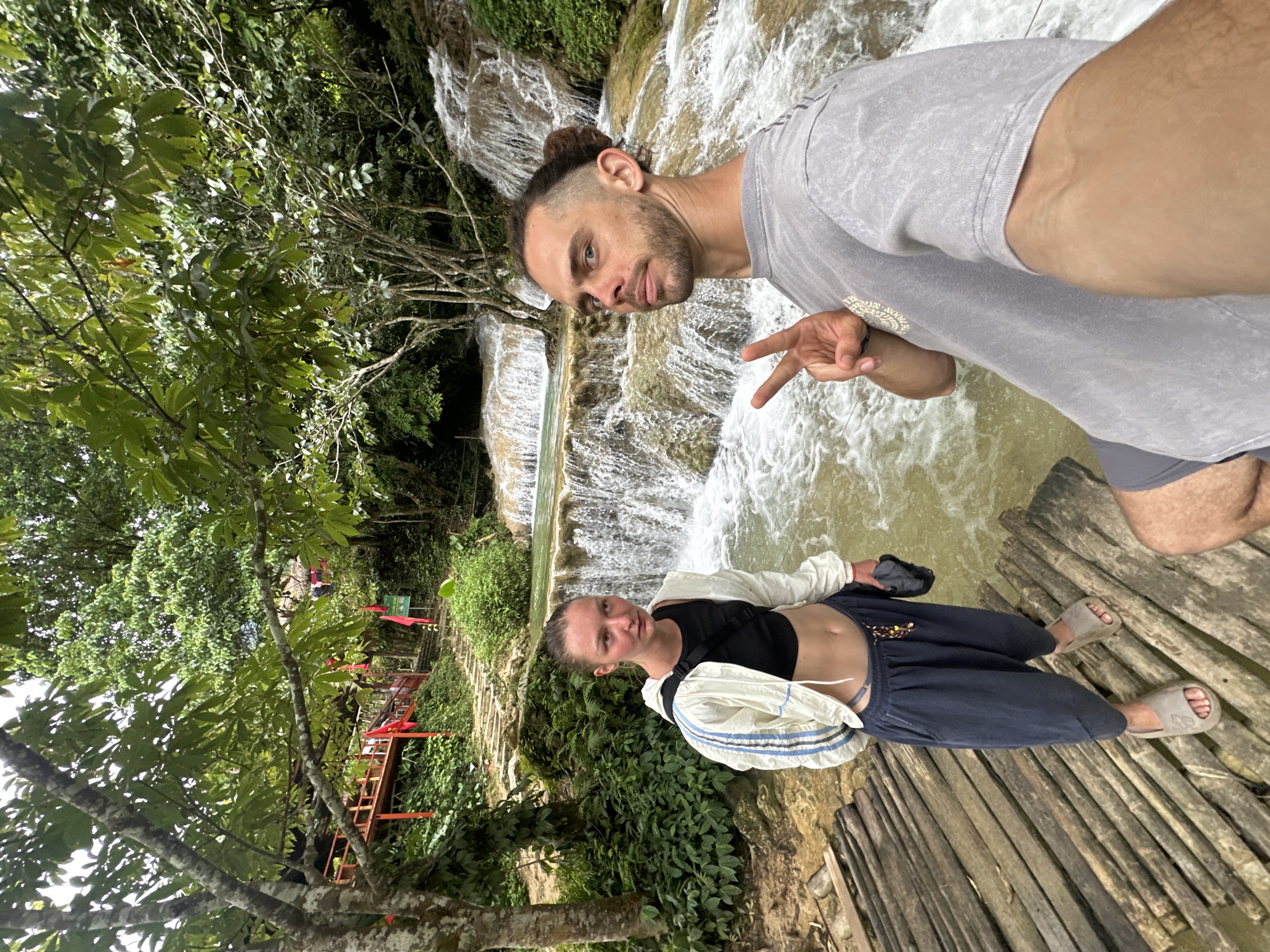 A man and a woman are standing on a wooden walkway in front of a tiered waterfall in a lush, green environment. The woman, on the left, wears a black crop top, a white jacket with blue and white stripes, and dark blue pants. She is looking to her left. The man, on the right, faces the camera, holding up two fingers in a peace sign. He wears a grey t-shirt and has his hair pulled back in a bun. The waterfall cascades over rocks, with trees and foliage surrounding the area. Red flags are visible on a pathway leading up the embankment behind the woman. The overall scene suggests an outdoor recreational setting, likely a tourist attraction in Cổ Lũng Commune, Vietnam.