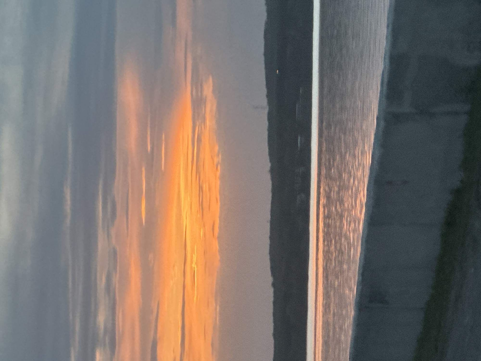 The image captures a sunset in Chaikovsky, Russia. The sky is a blend of blues and oranges, with the setting sun casting a warm glow. A body of water reflects the sunlight, creating shimmering streaks. A dark land mass separates the sky from the water.