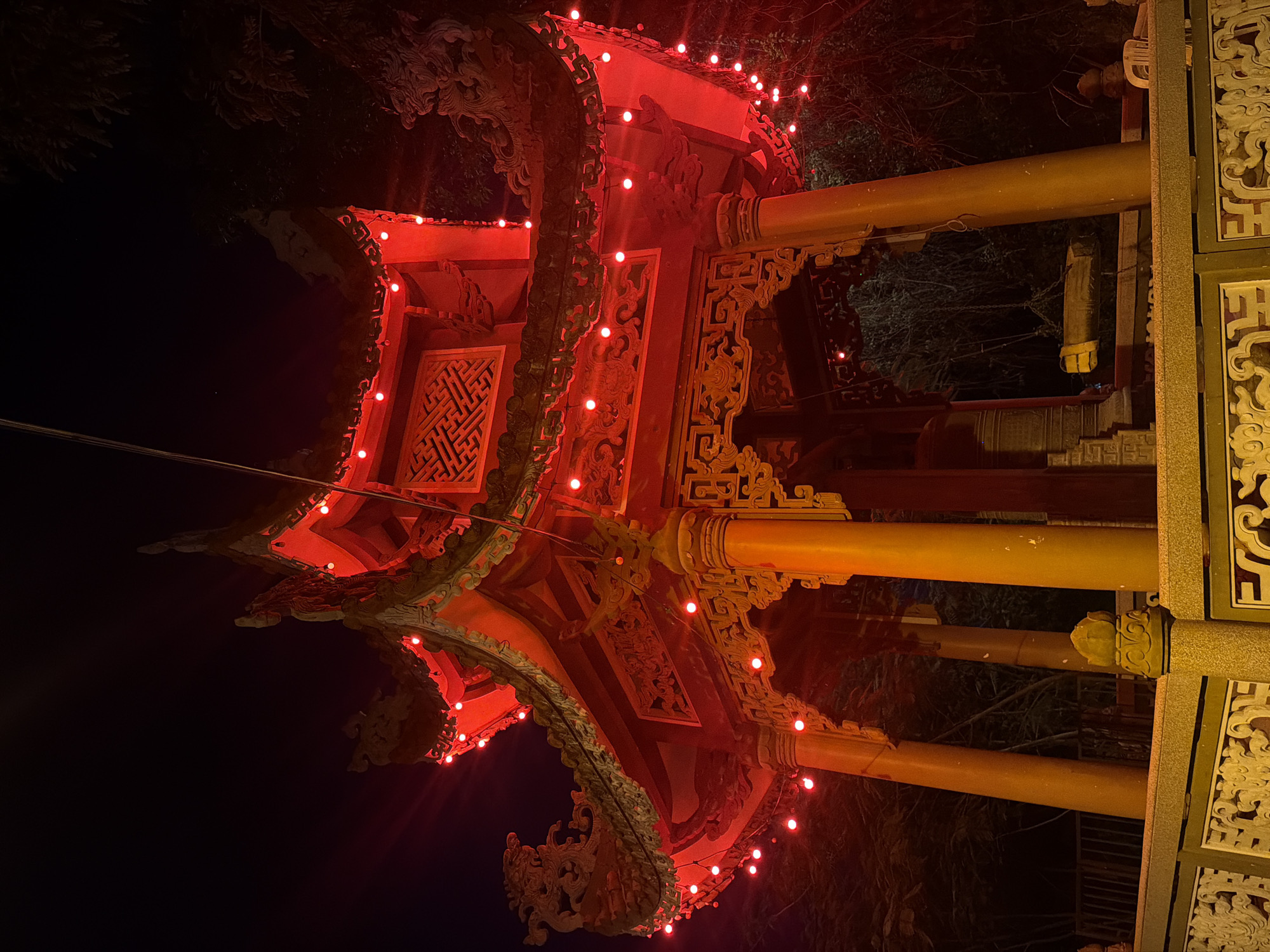 An ornate, multi-tiered pagoda, illuminated by numerous red string lights, stands prominently against a dark, nighttime sky. The pagoda's architecture features intricate carvings and decorative elements on its eaves and pillars. A large bell is visible hanging within the structure. The pagoda is situated within a landscaped area with trees visible in the background. The scene appears to be in a public or cultural space. This location is identified as a pagoda in Nha Trang, Vietnam.