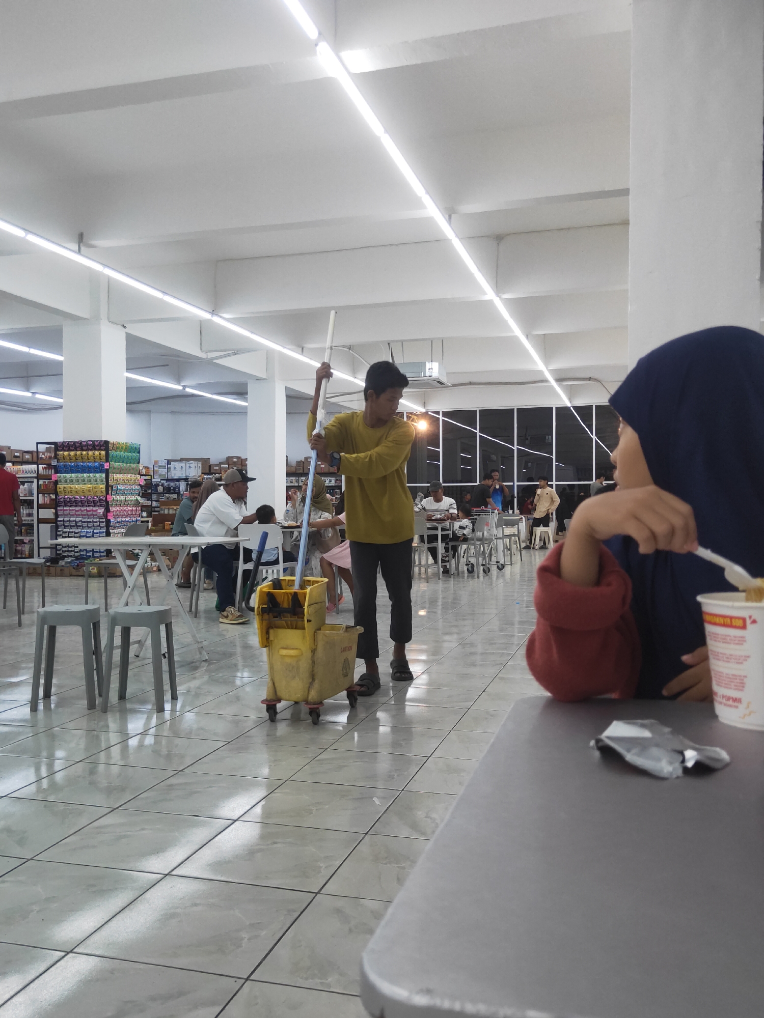 In the city of Sintang, Indonesia, inside what appears to be a spacious, brightly lit establishment with a tiled floor and high ceiling, a young man in a yellow shirt and dark pants is actively mopping. He wields a mop with a yellow bucket on wheels, the action suggesting a cleaning or maintenance task within the venue. In the foreground, a person wearing a navy blue hijab and a red top is seated at a table, partially obscuring the view. This person holds a white utensil, possibly a fork or spoon, and is looking to the left, their attention seemingly drawn to the mopping individual or other activities in the space. Other individuals are visible in the background, seated at tables and engaged in various activities, some appearing to be dining. Shelves stocked with products line the back walls, indicating this could be a supermarket, a cafeteria within a larger store, or a food court. The overall ambiance suggests a public or commercial space.