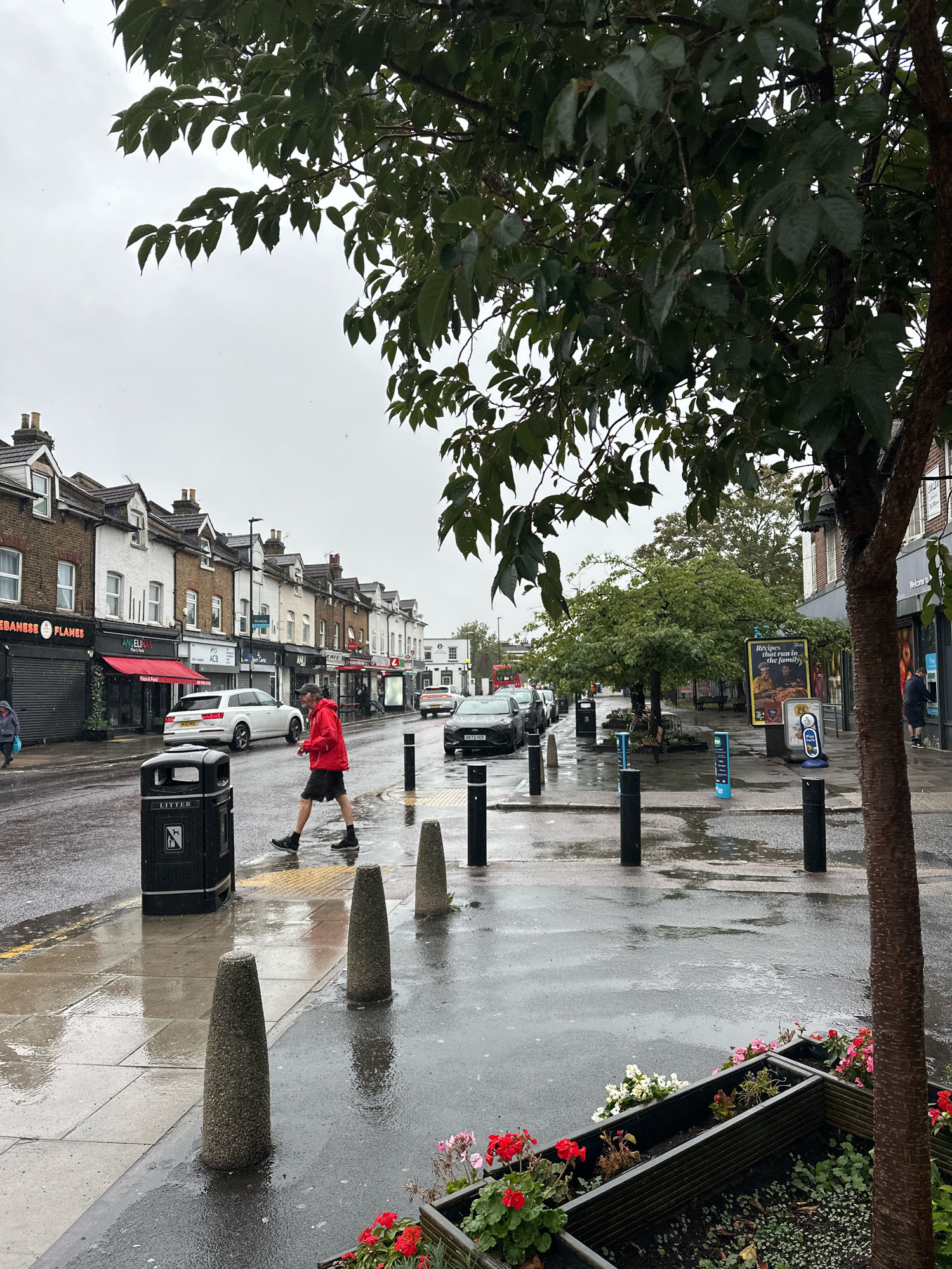 The image depicts a street scene on a rainy day in London, United Kingdom. A man wearing a red jacket and dark shorts is walking across a wet street, which reflects the overcast sky and the surrounding buildings. The street is lined with a row of shops with awnings, some displaying signs for businesses like "Lebanese Flames" and "Angelinas." Cars are parked along the street and a white Audi is driving away from the camera. A black litter bin is prominently positioned on the pavement. There are bollards along the sidewalk and a planter box with red and pink flowers in the foreground. The overall atmosphere is damp and muted due to the weather.