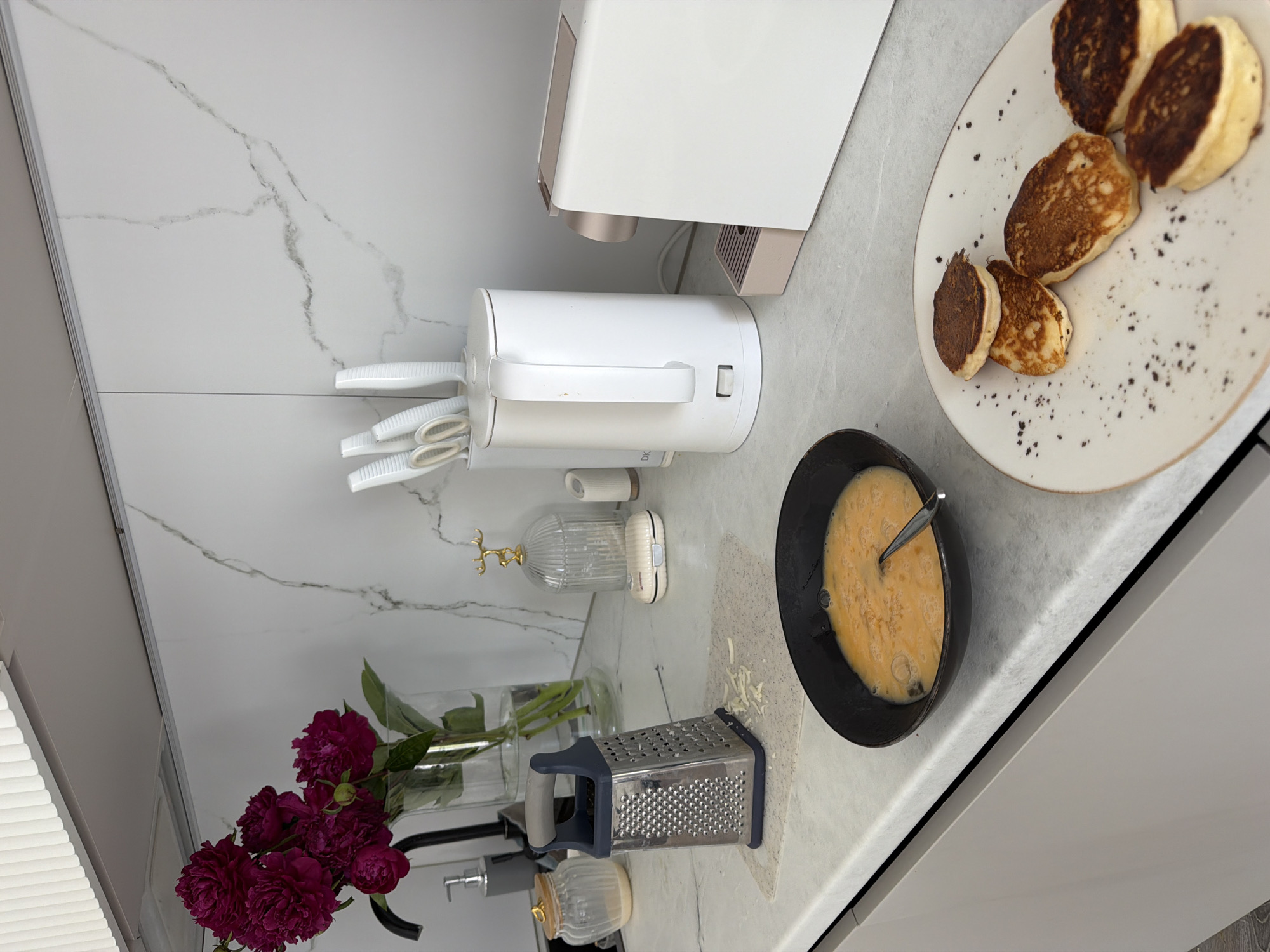A well-lit, modern kitchen counter in Malinovka, Russia, showcases breakfast preparations. A bouquet of dark pink peonies in a glass vase sits to the left, beside a matte grey soap dispenser and a clear glass jar with a wooden lid. A silver box grater with shredded cheese on a light grey mat is central. A black bowl filled with a whisked egg mixture and a spoon rests in front of the grater. To the right, a white electric kettle stands next to a white and rose gold coffee machine. A white speckled plate holds five small, golden-brown pancakes, some dusted with dark specks. The background features a white marbled backsplash.