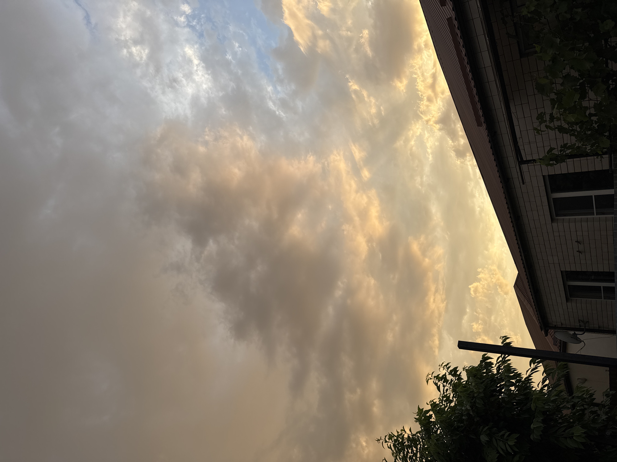 The image depicts a cloudy sky with warm, sunset hues over a residential area. In the foreground, the dark green foliage of a tree is visible on the left. A dark, vertical post stands prominently near the center. To the right, the red-tiled roof of a building is seen, with a brick wall beneath it. The building appears to be a house with windows. A satellite dish is mounted on the wall of the house. The sky is filled with a mix of white, grey, and orange clouds, suggesting either dawn or dusk. The scene appears to be located in Mikhaylovsk, Russia.