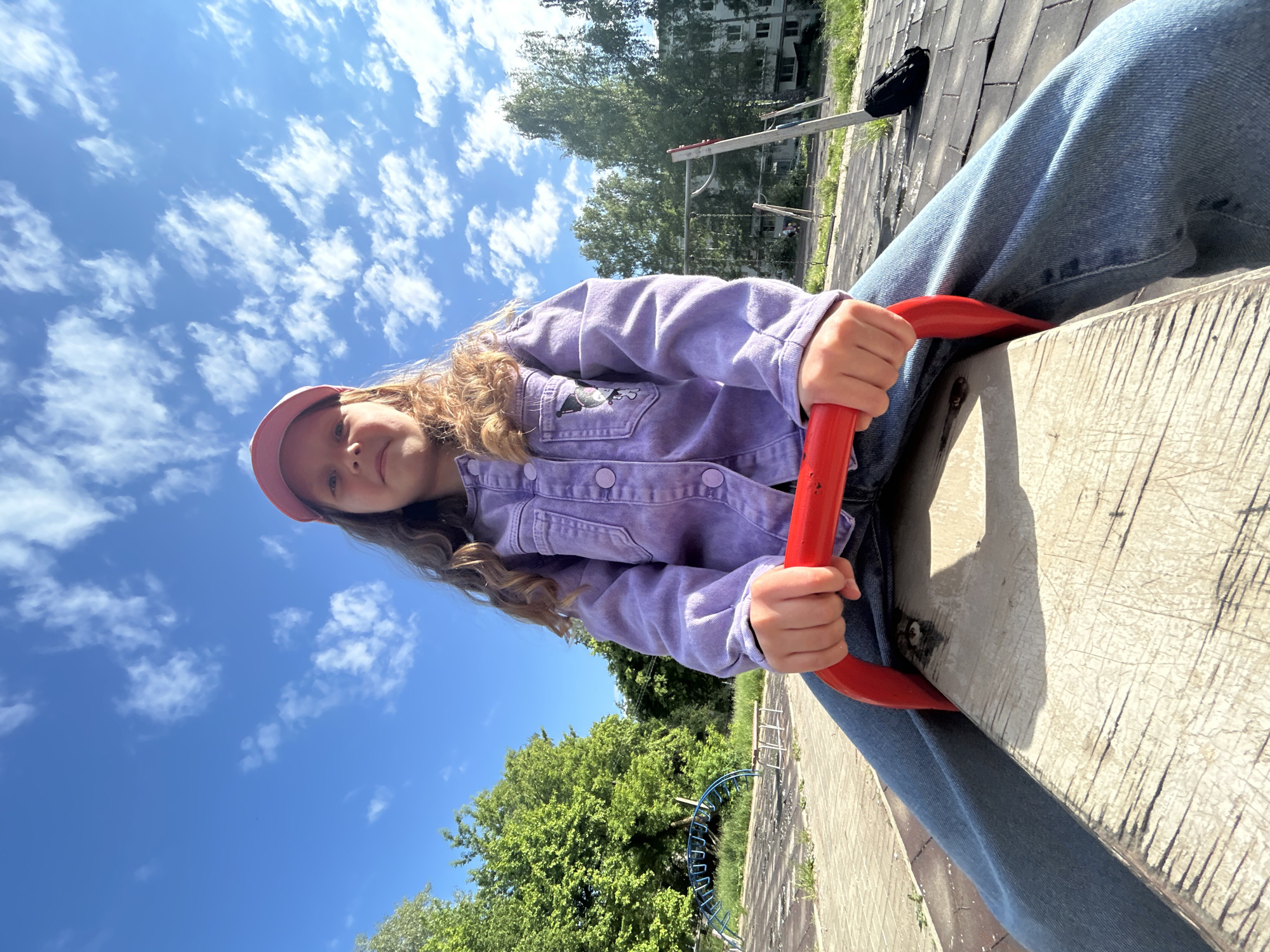 A young girl is prominently featured in a low-angle outdoor shot, seated on a seesaw and looking directly at the camera with a gentle smile. She has long, wavy light brown hair and is wearing a light purple denim jacket with a cartoon patch on one pocket, blue jeans, and a pink baseball cap. Her hands are gripping a red handle, which is part of the light-colored seesaw seat.
The scene is set in what appears to be a playground or recreational area in Chaikovsky, Russia, under a bright blue sky filled with scattered white clouds, indicating a sunny day, likely midday or early afternoon. In the background, lush green trees are abundant, and a multi-story residential building, possibly an apartment block of an older architectural style, is visible. Another blue metal playground structure can be partially seen through the trees. The ground around the playground consists of both paved sections, possibly tiles, and natural grassy or dirt areas. A black backpack rests on a paved surface in the distance. The overall atmosphere of the image is cheerful and captures a moment of outdoor play. No visible text is discernable in the image.