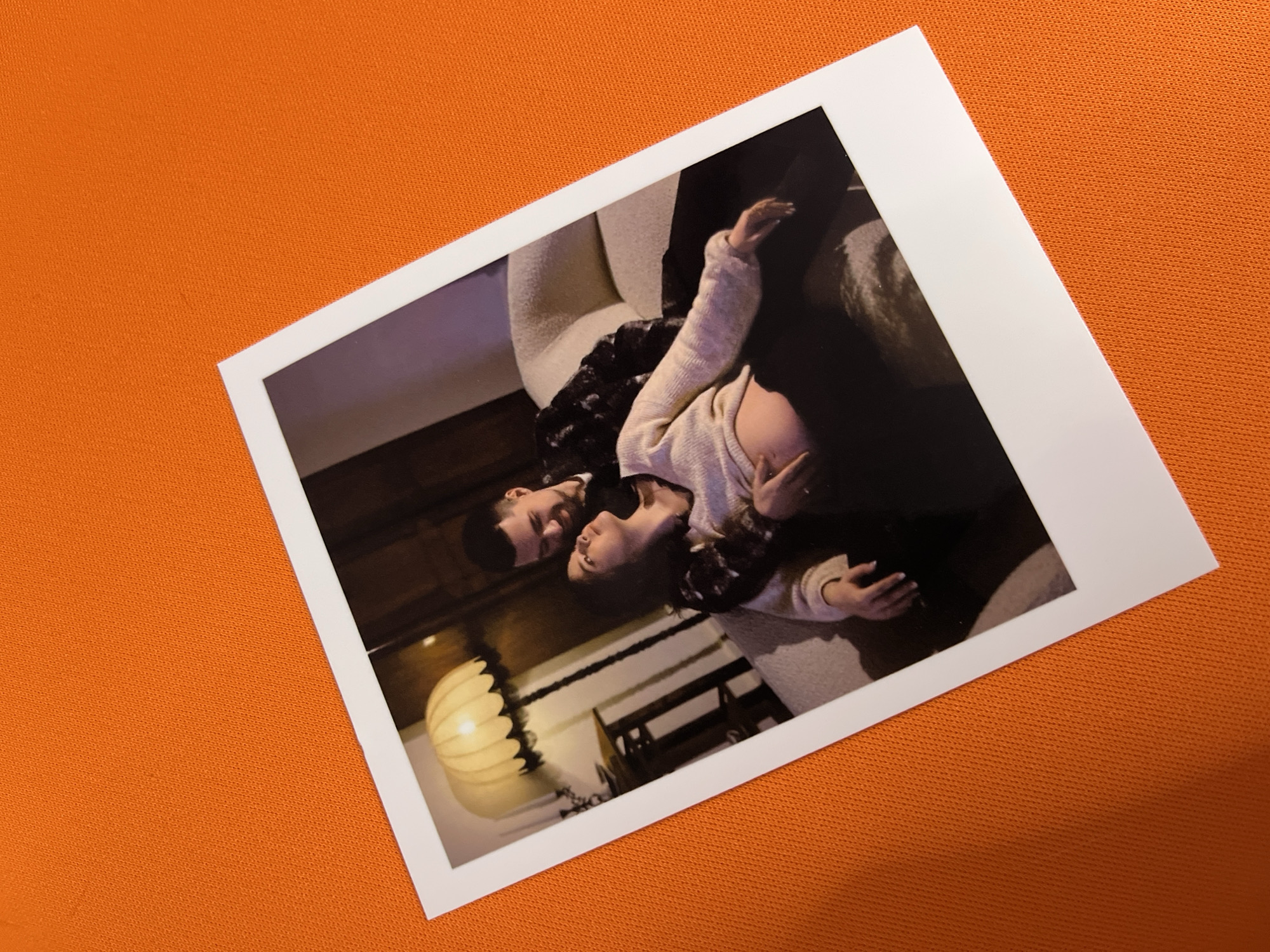 An instant film photograph, oriented diagonally, rests on a bright orange, textured fabric surface. The print features a man and a woman in a close pose, lying down and gazing at each other. The woman is positioned closer to the viewer, wearing a light-colored, possibly cream, knitted top, with her dark hair partially covering her shoulders. The man, behind her, is dressed in a dark patterned top. They are situated on what appears to be a light-colored, plush sofa or armchair, creating a cozy indoor scene.
The lighting in the photograph is dim, suggesting an evening or night setting, with artificial light emanating from a distinctive, ribbed, gourd-shaped lamp visible on the left side. Wooden furniture or paneling can be seen in the background, adding to the warm, intimate atmosphere. Both individuals appear relaxed and affectionate, looking intently at each other. The white border of the photograph is prominent, characteristic of instant prints. No text is visible in the image. The scene is set in Shushary, Russia.