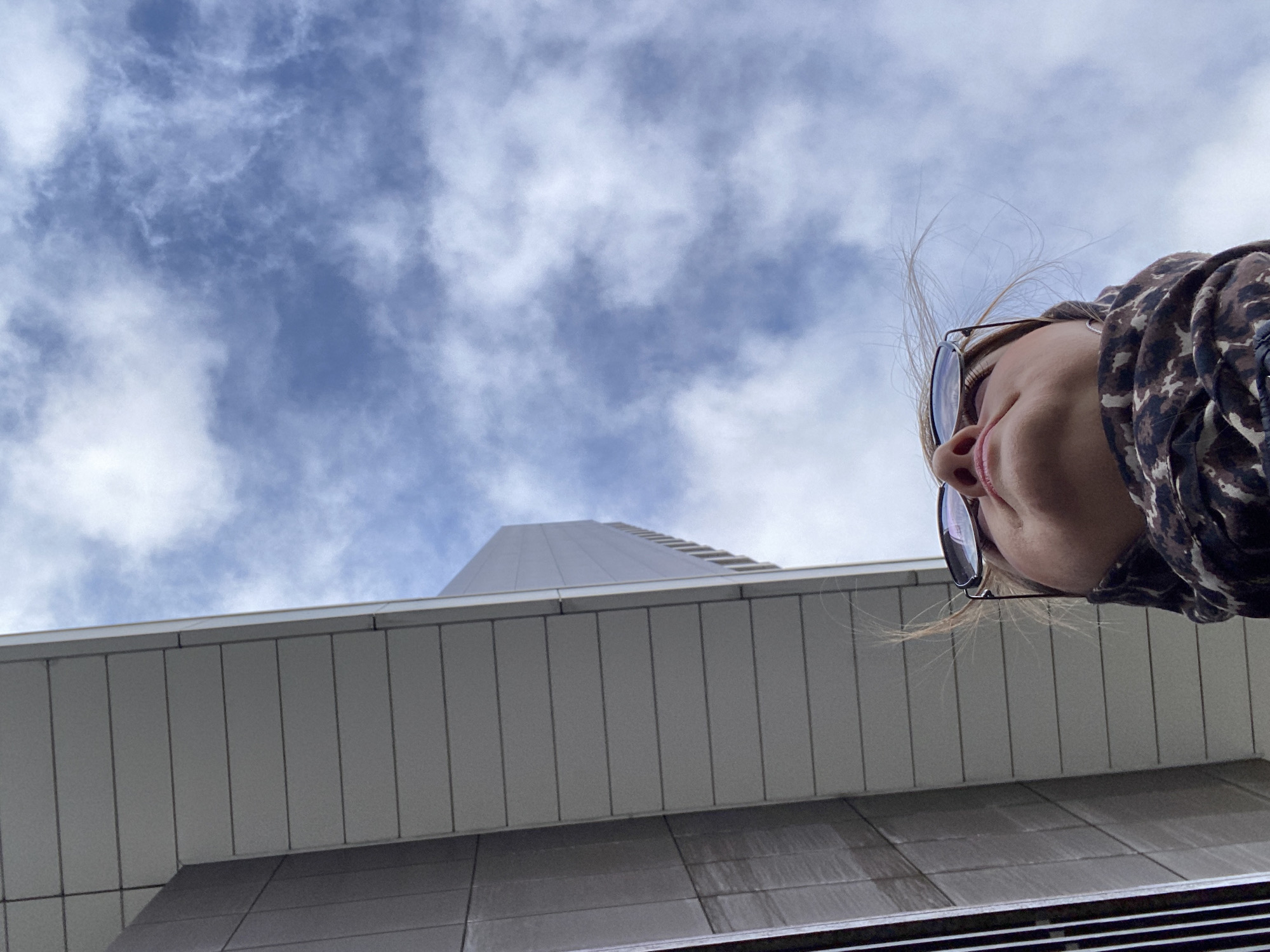 The image is a low-angle shot looking up. In the foreground is the lower portion of a person's face, including the nose, mouth, and chin, wearing glasses and a patterned hoodie. Behind the person is a modern building facade composed of gray, vertically oriented panels. Above the building, the image shows a tall, white building extending toward a cloudy, blue sky. Although the exact location is unclear, it is within the city of Yekaterinburg, Russia.