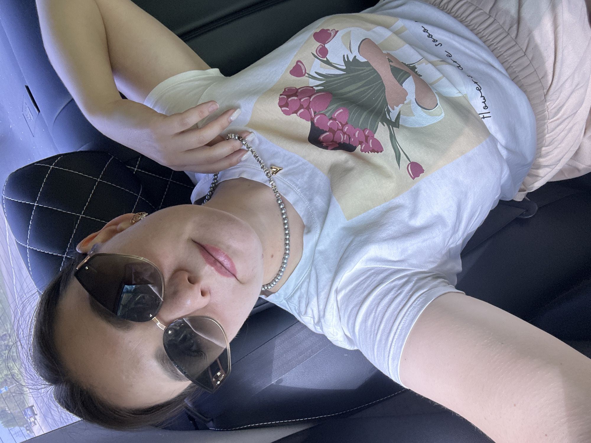 A young woman reclines in the back seat of a car. She wears sunglasses, a white t-shirt with a floral graphic, beige shorts, and a silver beaded necklace. Part of a black leather seat with white stitching is visible. The photograph appears to be taken from a first-person perspective. Based on the slight blurriness of the image, the location is a moving vehicle. The partial reflection of buildings and trees outside the car window points to an urban setting. While it is impossible to pinpoint the location within Chaikovsky, Russia with certainty from the image alone, the details suggest the photo was taken while traveling through the city.