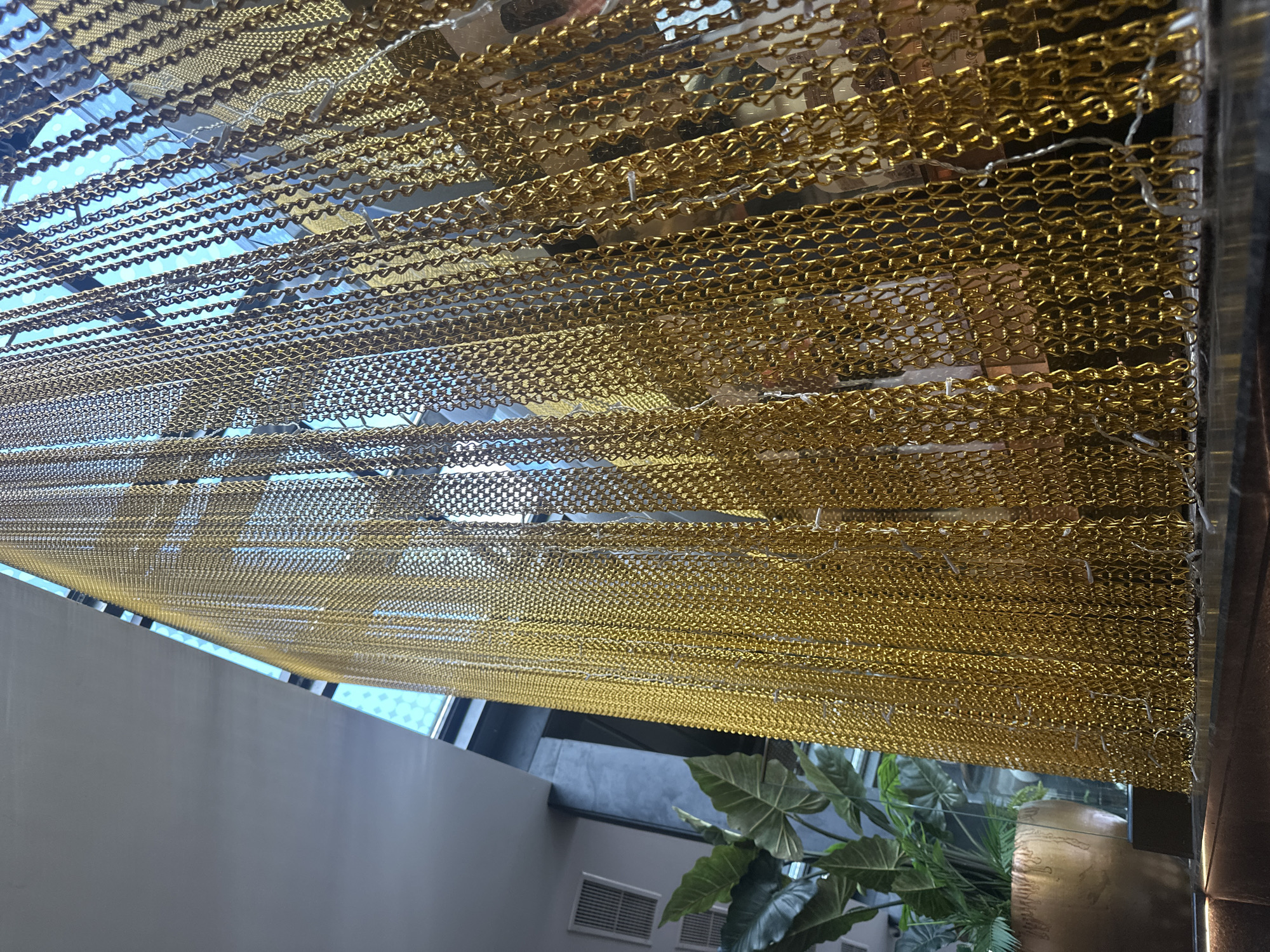 An interior shot shows a cascade of gold chains hanging vertically, creating a textured curtain. The chains are tightly packed, forming a shimmering, reflective surface that catches the light. Behind the chain curtain, a bright blue sky with some geometric patterns is visible through a large window or skylight. To the left of the chains, a large potted plant with broad green leaves adds a touch of nature to the scene. The overall impression is one of modern, possibly luxurious, interior design.