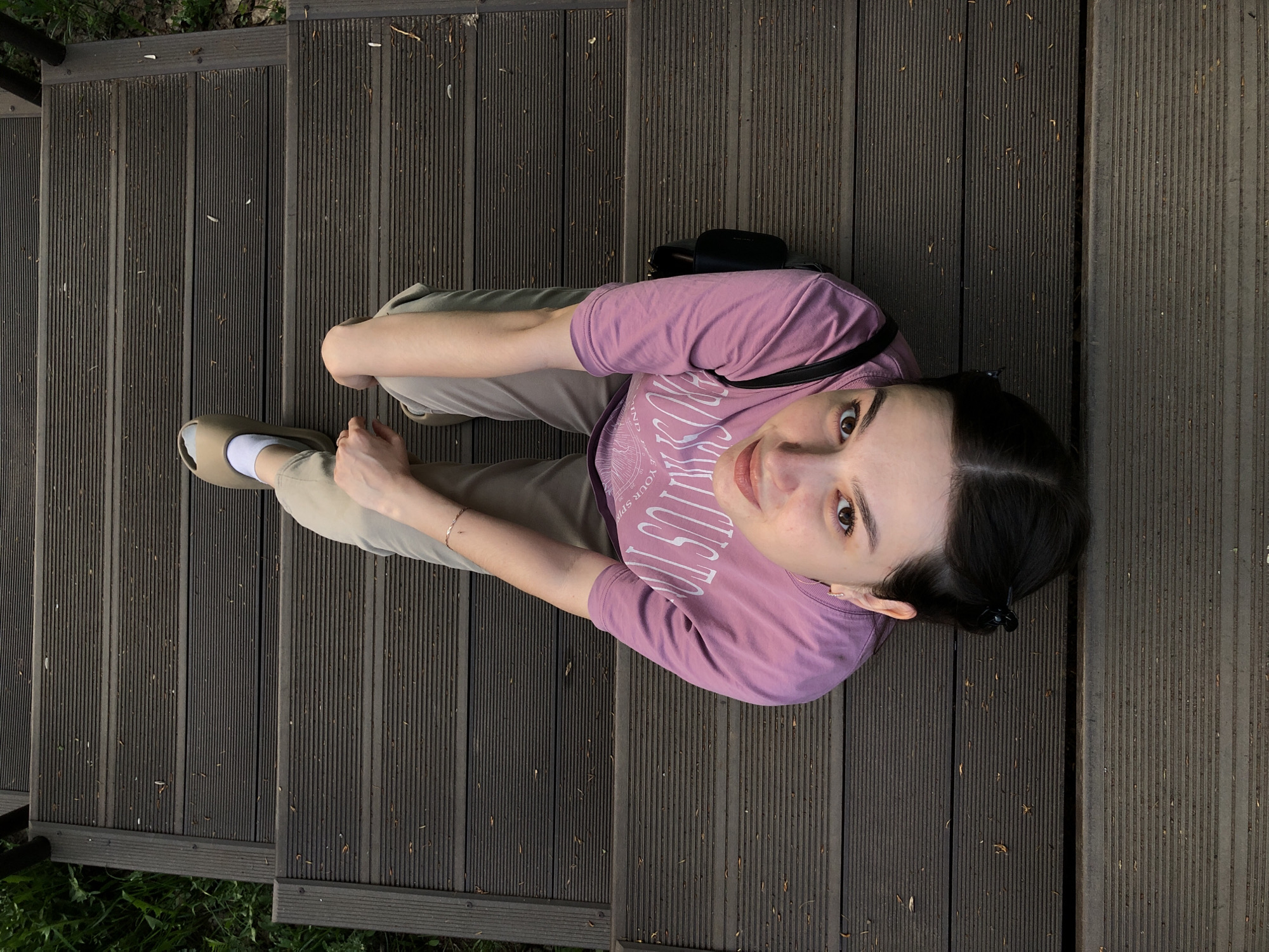 A high-angle shot depicts a young woman sitting on wooden stairs outdoors. She is wearing a pink t-shirt and light brown pants with light brown shoes and white socks. She has dark hair pulled back with a hair clip. A black purse is visible behind her. The stairs are made of dark wood planks with thin grooves running along them. Green foliage is visible at the bottom of the frame. The photo was taken in the city of Chaikovsky, Russia.