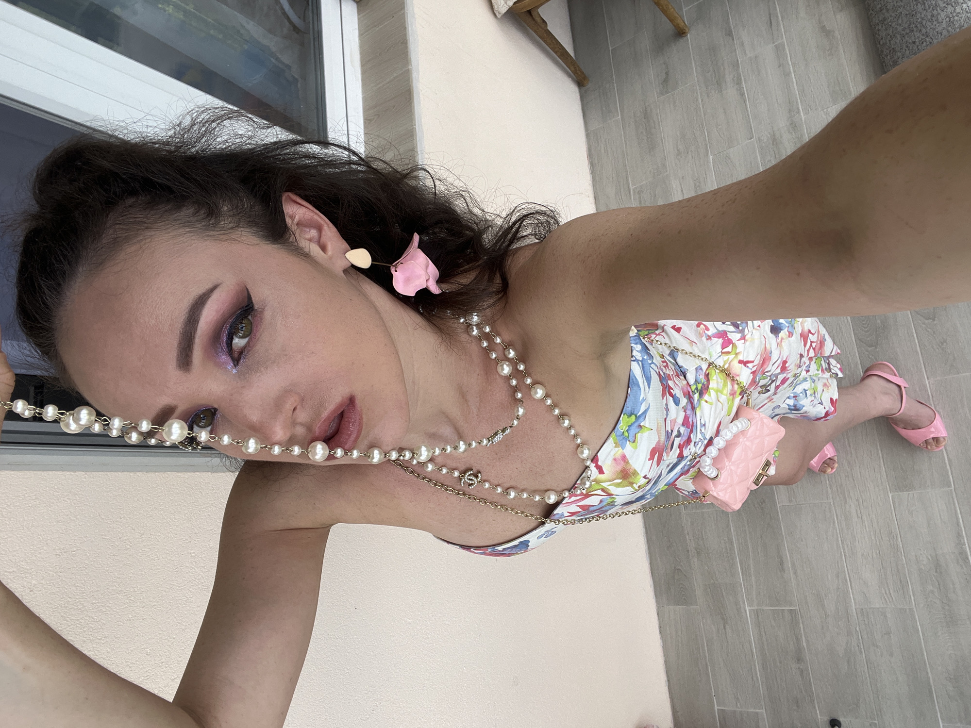 A woman is standing outside, with her right arm extended above her head, holding a string of pearls. She is looking down at the camera. She is wearing a floral print dress, pink pearl-embellished handbag, and pink sandals. She has on dramatic eye makeup, including pink eyeshadow and black eyeliner. She also has a pink petal-shaped earring in her left ear. The background appears to be a building with a light-colored wall and a large window. The ground is tiled. This image was not taken in Kazan, Russia.