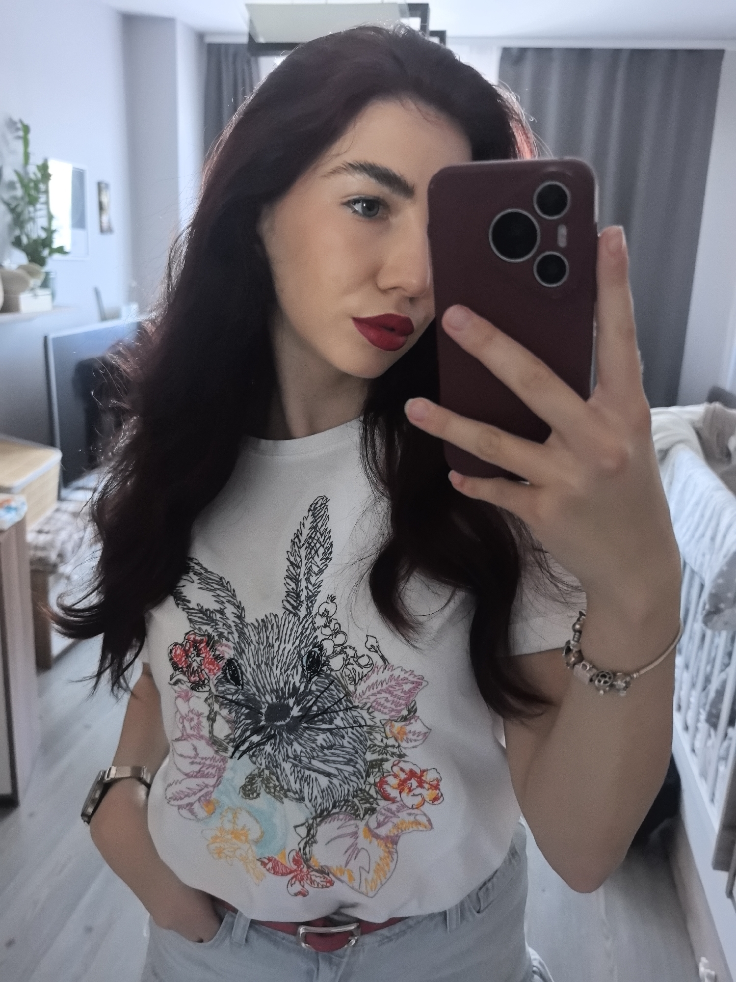 A young woman takes a selfie in an indoor setting that appears to be a home or apartment. The location is likely Stara Zagora, Bulgaria, due to the user's location. The woman holds a smartphone with a burgundy case partially obscuring her face. She has dark hair, fair skin, and red lipstick. She wears a white t-shirt with a rabbit and floral design, light blue denim jeans, a red belt, a bracelet, and a wristwatch. The background shows a room with light-colored flooring, white walls, a crib, and a dresser. A glimpse of a television and decorative plants is visible.