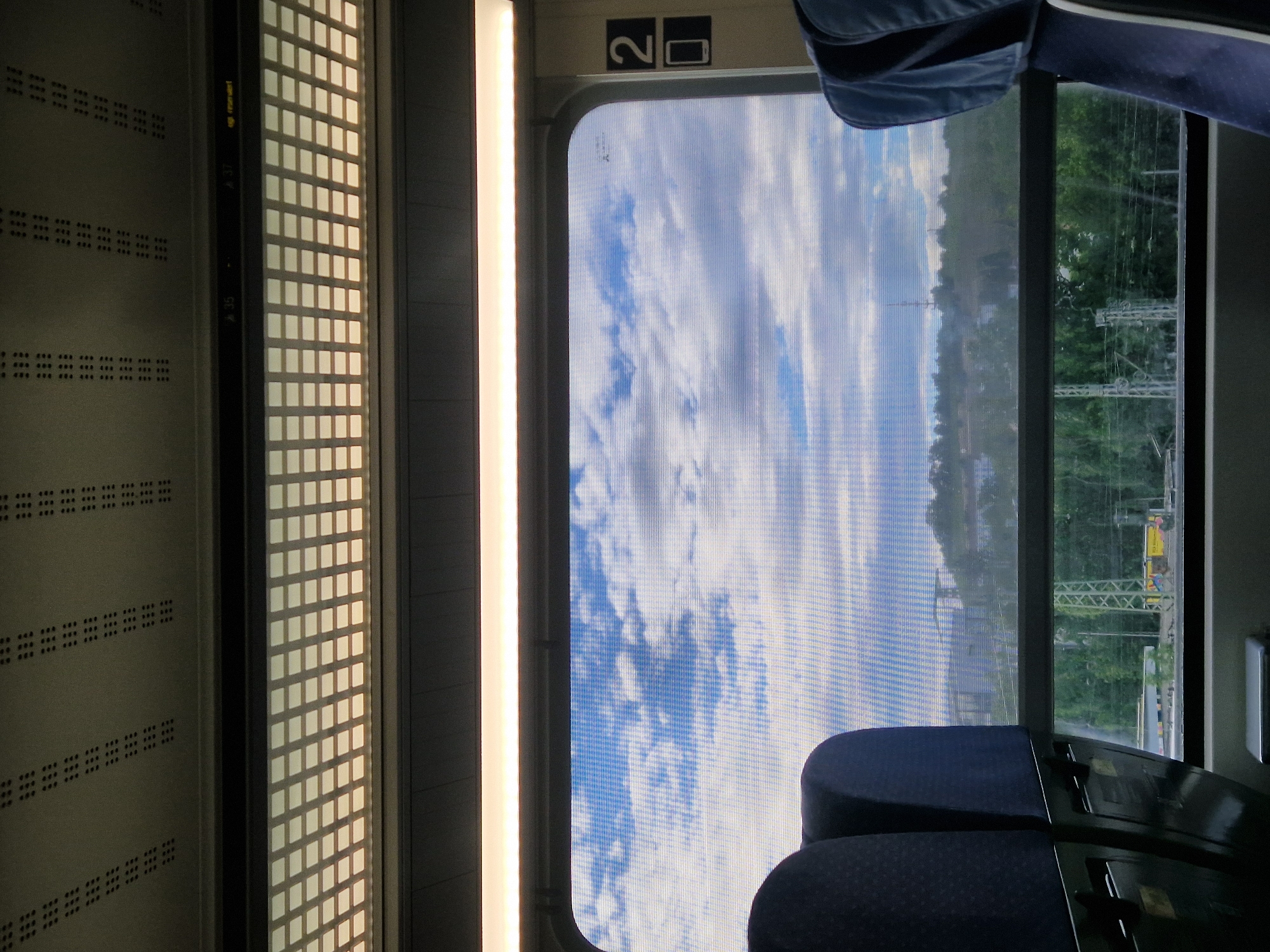 The image depicts the interior of a train car, with a view out of a window. On the left side of the frame, the train's interior wall is visible, featuring a patterned design of small squares and rows of what appear to be luggage racks. A vertical strip of light runs along the right edge of this interior wall.
The dominant feature is the train window, which looks out onto a bright, cloudy sky and a landscape of trees. The sky is a mix of blue and white clouds, with a textured, possibly netted, quality to the window itself, causing the view to appear slightly distorted or pixelated. The landscape outside consists of lush green trees lining a hillside. In the distance, train tracks and some infrastructure, including what looks like a yellow vehicle, are visible.
At the top of the window frame, a sign with the number "2" and a symbol that resembles a luggage icon is present. The seats of the train are visible in the foreground and bottom of the frame, upholstered in a dark blue fabric.
The overall scene suggests a daytime journey, likely in fair weather given the bright sky. The "Stuttgart, Germany" location context implies this is a train operating within or near that city. There is no clear indication of people or specific activities taking place, other than the implied act of traveling. The lighting suggests it is daytime, possibly late morning or afternoon.