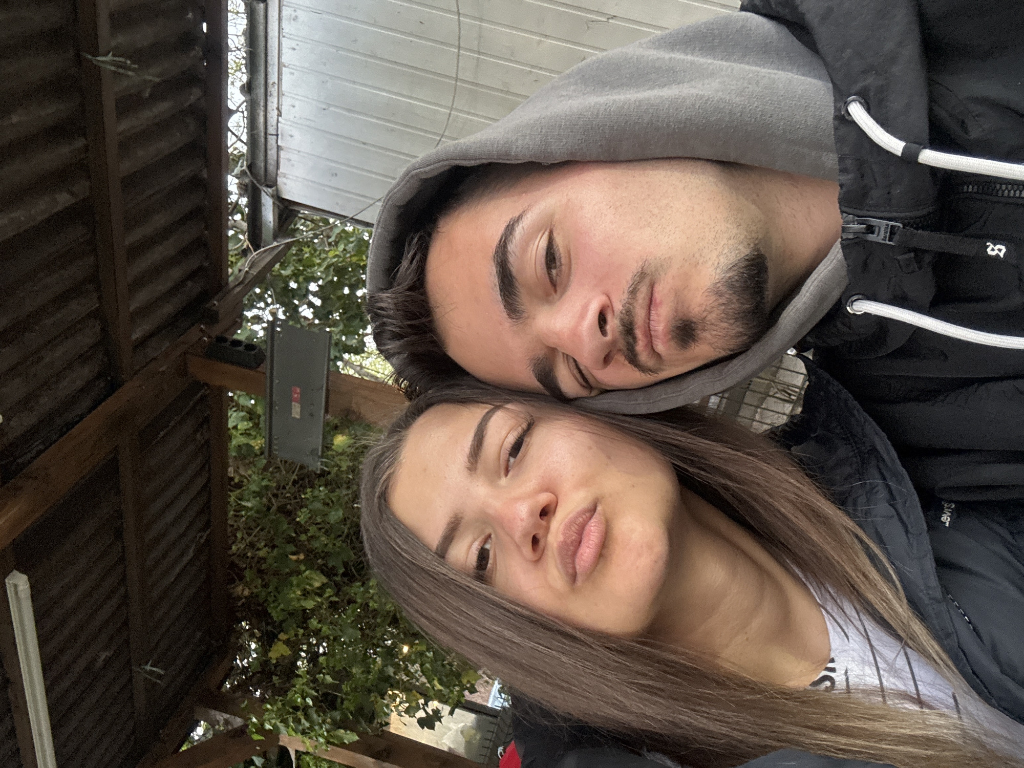 This media file captures a close-up, rotated selfie-style portrait of two young individuals, a male and a female, taken outdoors in what appears to be Sofia, Bulgaria. The image is rotated 90 degrees clockwise, giving the impression that the subjects are resting their heads together, looking up at the camera.
The male, positioned on the right side of the frame (when viewed upright), wears a grey hooded sweatshirt under a black puffer jacket. He has dark hair, prominent eyebrows, and a neatly trimmed beard and mustache. His eyes are slightly narrowed or downcast, conveying a relaxed or contemplative mood. White drawstrings are visible on his black jacket, along with a metallic zipper and a small white graphic that resembles an abstract symbol or letters like "BS" or "B8".
The female, to the left, has long, straight brown hair and appears to be wearing a white top beneath a black jacket. She has a distinctive pouting expression, suggesting playfulness or an intentional pose. A partial white logo, possibly "LEVI'S," is visible on her black jacket near her shoulder.
The setting appears to be an outdoor, semi-sheltered area, possibly a patio, balcony, or a pergola. Wooden slatted structures form parts of the background, along with lush green foliage from trees or bushes, indicating a natural environment. A light-colored corrugated structure or roof and a grey electrical box mounted on the wooden framework are also visible in the background. The lighting is bright and diffused, suggesting it is daytime, possibly under an overcast sky or in indirect sunlight. The overall emotion conveyed is one of casual intimacy and youth.