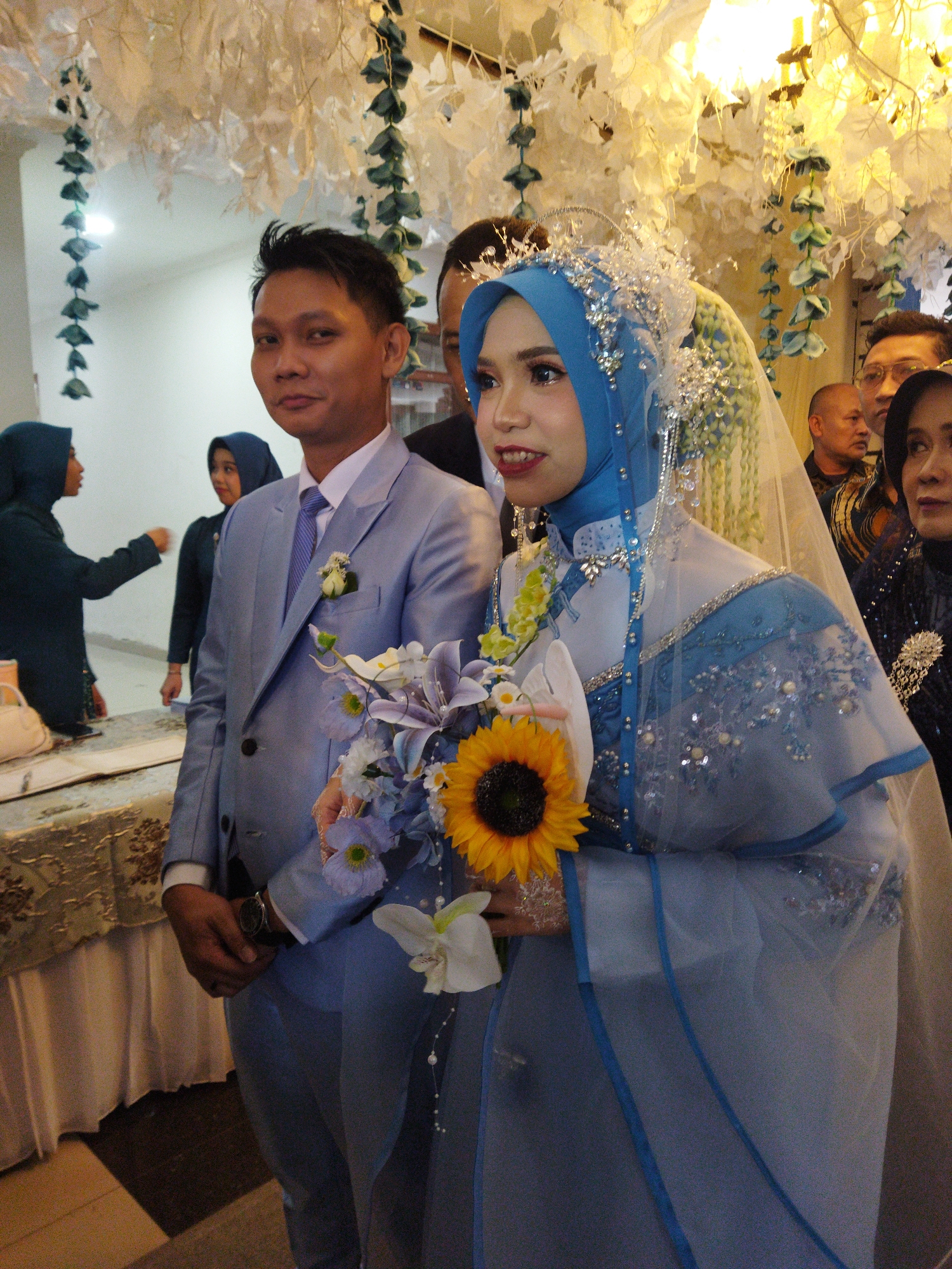 A medium shot captures a wedding ceremony in Semarang, Indonesia, with a newlywed couple in the foreground. The groom, on the left, wears a light blue suit with a white shirt and purple tie, a boutonniere on his lapel. He holds a bouquet of flowers, including a sunflower, in his right hand. The bride, to his right, is adorned in a blue hijab and a matching wedding gown with intricate beadwork and embroidery. A veil with sparkling embellishments covers her head. She is also holding the bouquet. Behind them, several guests are visible, some partially obscured. In the background, the venue is decorated with white fabric and hanging greenery. To the far left, a woman in a teal hijab and dress is partially visible, and a table with a white cloth and what appears to be a purse is set up. The lighting suggests an indoor event.