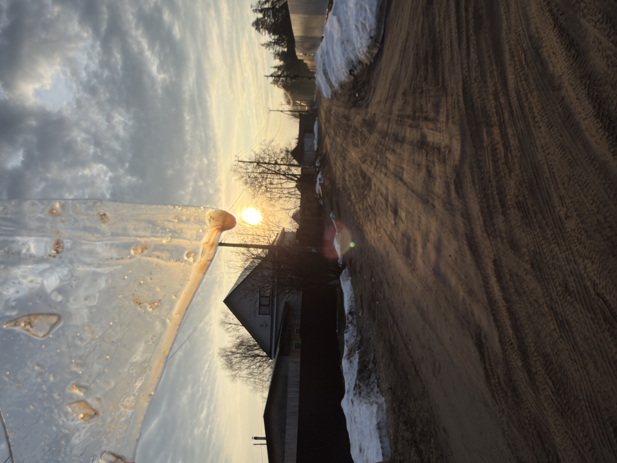 The image depicts a dirt road with tire tracks, located in what appears to be a rural or semi-rural area near Chaikovsky, Russia. Remnants of snow are visible along the roadside. A residential building with a pointed roof stands to the side of the road. The sky is overcast with a setting sun visible through the trees. A translucent object, possibly glass or ice, is in the foreground, obscuring part of the scene.
