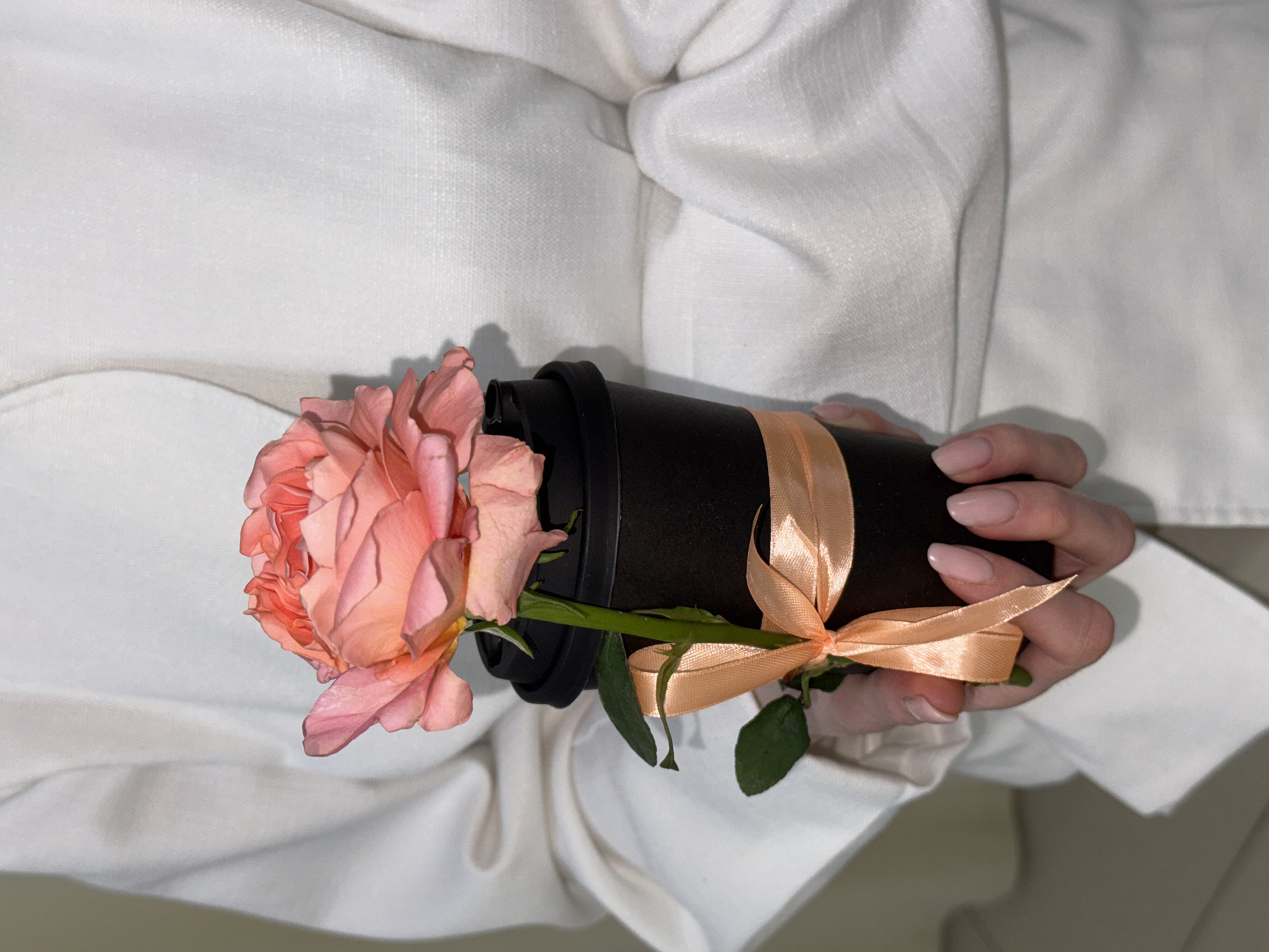 A close-up shot depicts a person holding a black reusable coffee cup, adorned with a peach-colored ribbon, in which a peach-colored rose is placed. The person is wearing a white linen shirt. The scene's location is Chaikovsky, Russia.