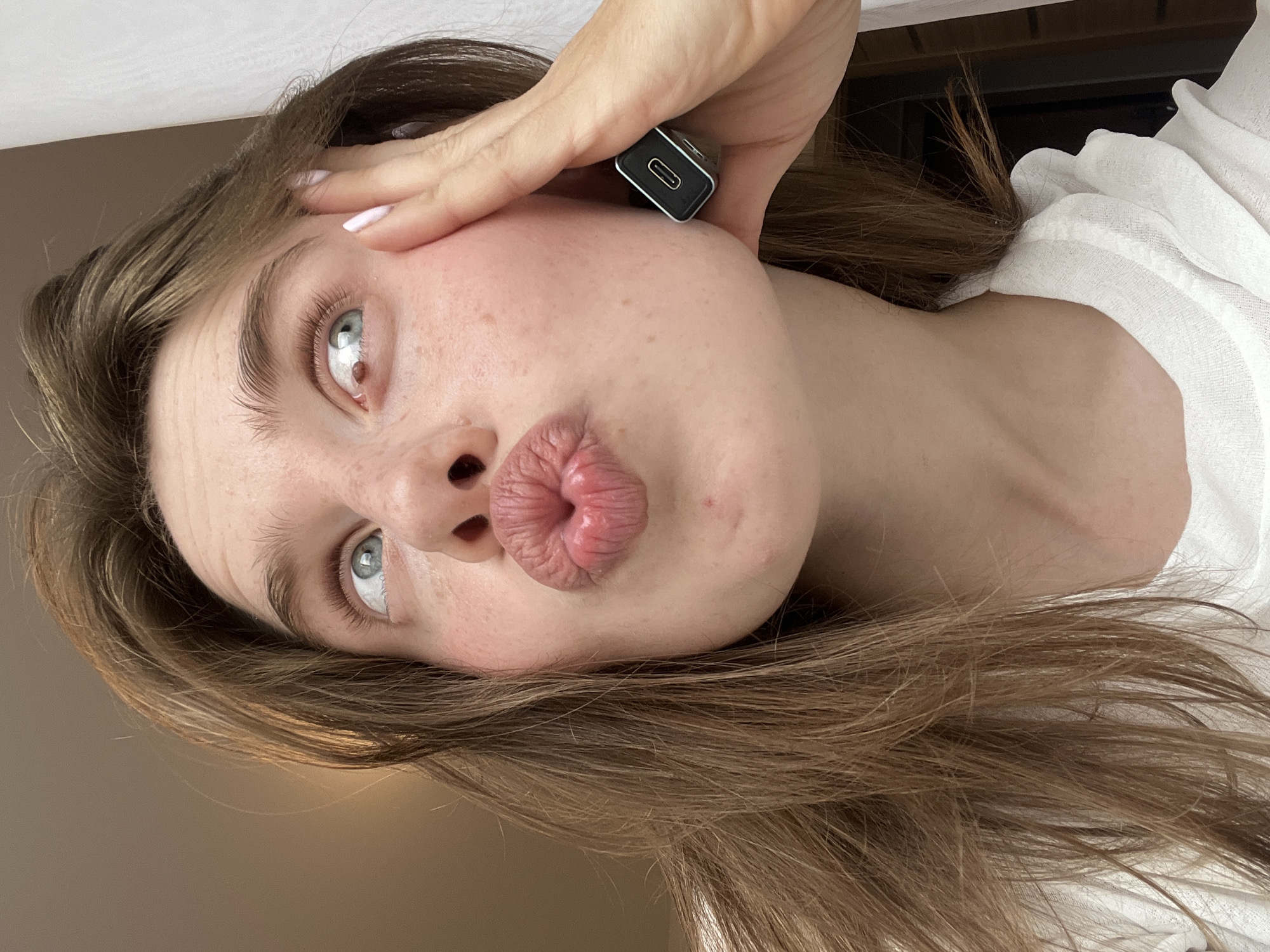 A close-up, waist-up, selfie-style shot shows a young woman with light brown hair, blue eyes, and fair skin with freckles. She is looking up and to her right with her lips pursed. Her right hand is on her cheek, and she is holding a small black rectangular object with a USB-C port between her fingers. The background is out of focus and appears to be indoors with a white curtain on the right side. The image is well-lit, likely with natural light. There is no indication of the location being in Chaikovsky, Russia, as there are no identifiable landmarks or elements that suggest this specific city.