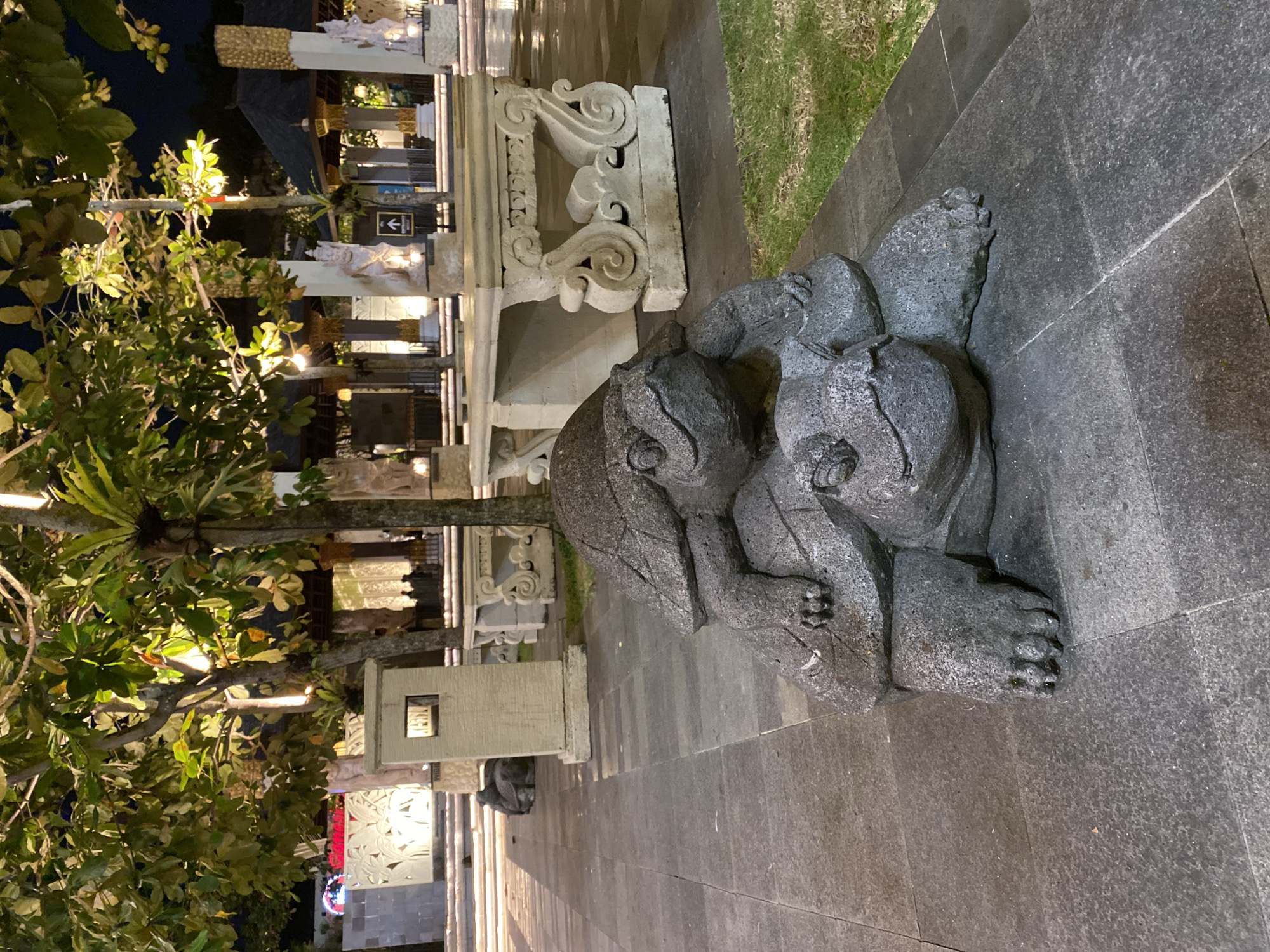 In Kuta Selatan, Indonesia, a stone sculpture of two turtles stacked on top of each other is prominently displayed on a stone-tiled pathway at night. The larger turtle forms the base, with its head facing forward and its legs tucked beneath it. The smaller turtle rests on the back of the larger one, its head also facing forward, appearing to look over the larger turtle's shell. Both sculptures are detailed, showcasing textured shells and distinct facial features.
Behind the turtle sculpture, a paved area leads to an outdoor seating area with ornate stone benches and tables. Trees with lush green foliage are visible, their leaves illuminated by ambient lighting, casting a warm glow. In the background, there are traditional Indonesian-style structures with thatched roofs and decorative pillars, suggesting a cultural or resort setting. Several people are faintly visible in the distance, silhouetted against the illuminated buildings, and an "ENTRANCE" sign with an arrow is posted on a pillar. The ground is wet and reflects the lights, indicating recent rain. The overall scene conveys a tranquil, possibly tourist-oriented, outdoor environment at dusk or night.