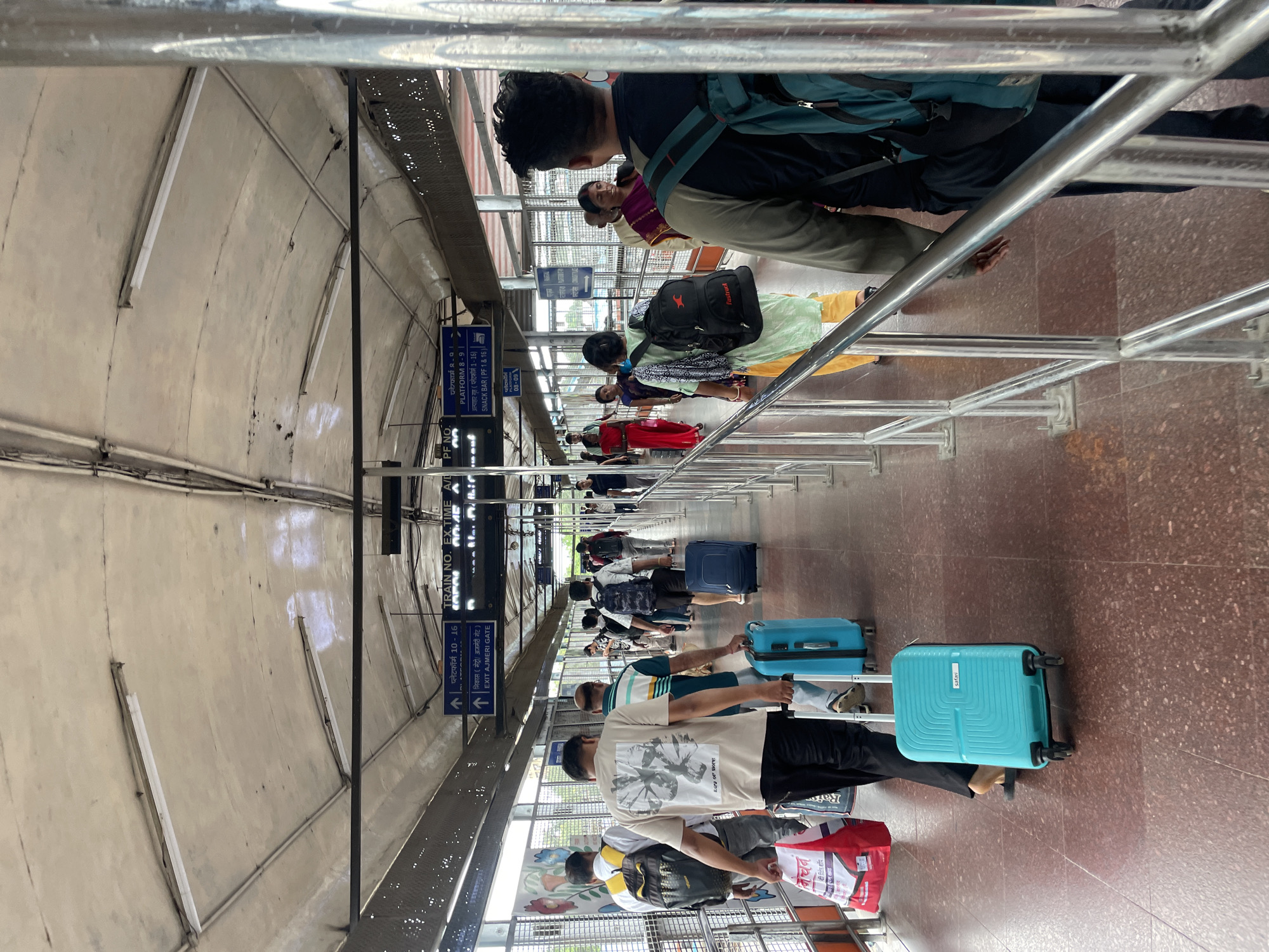 The image depicts an interior view of a busy, covered pedestrian walkway, likely at a train station in New Delhi, India. The walkway is lined with metal railings and a tiled floor. Overhead, fluorescent lights illuminate the scene, and signs indicating platform numbers and exits are visible. A diverse group of people, dressed in casual clothing and some in traditional Indian attire, are seen walking along the walkway, many carrying luggage, including suitcases, duffel bags, and shopping bags. The overall atmosphere suggests a sense of movement and transit, typical of a public transportation hub.
