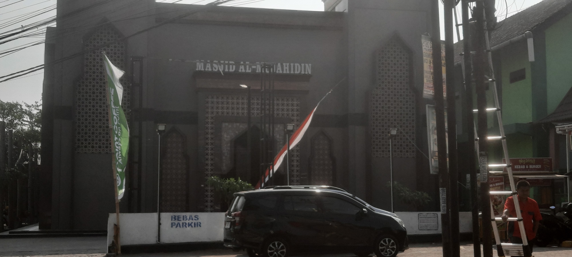 In Jeblog, Indonesia, a mosque named "MASJID AL-MUJAHIDIN" is visible, characterized by its dark facade and intricate patterned windows. A green and white flag, bearing the text "POINT COFFEE Always on Point," stands to the left of the mosque entrance, while an Indonesian flag with red and white stripes is positioned in front of it. A black minivan is parked in front of a white barrier with the words "BEBAS PARKIR" (Free Parking) written on it. To the right of the scene, a man in an orange shirt stands near a ladder leaning against a pole. Behind him, a building with a green facade houses a food stall advertising "Nyamil KEBAB & BURGER." The environment appears to be an urban street with overhead electrical wires, indicative of a bustling city area.
