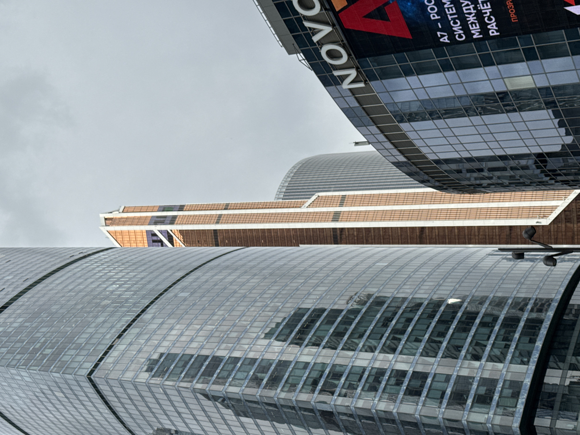 The image depicts the architectural details of buildings in Khimki, Russia, seen from a low angle. The most prominent features are the glass-clad surfaces of modern skyscrapers, reflecting the sky. One building displays a large advertisement with the word "NOVO" visible. In the background is a building with horizontal brown stripes and a curved roof. The sky is overcast. The overall impression is of a modern urban landscape.