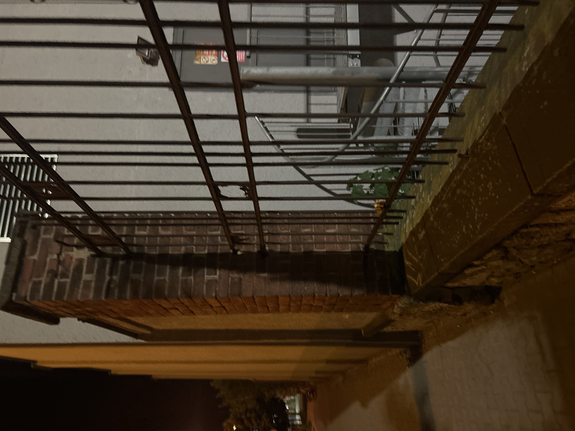 The image depicts a nighttime scene outdoors in Pilsen, Czechia. In the foreground, a paved sidewalk runs alongside a textured, yellow building on the left. Adjacent to the building is a brick pillar and a dark metal fence. Behind the fence, a spiral staircase is visible, leading upwards. The lighting suggests it is dark, with artificial light casting shadows and illuminating parts of the scene. The overall impression is that of an urban environment at night, possibly near an entryway or a service area.