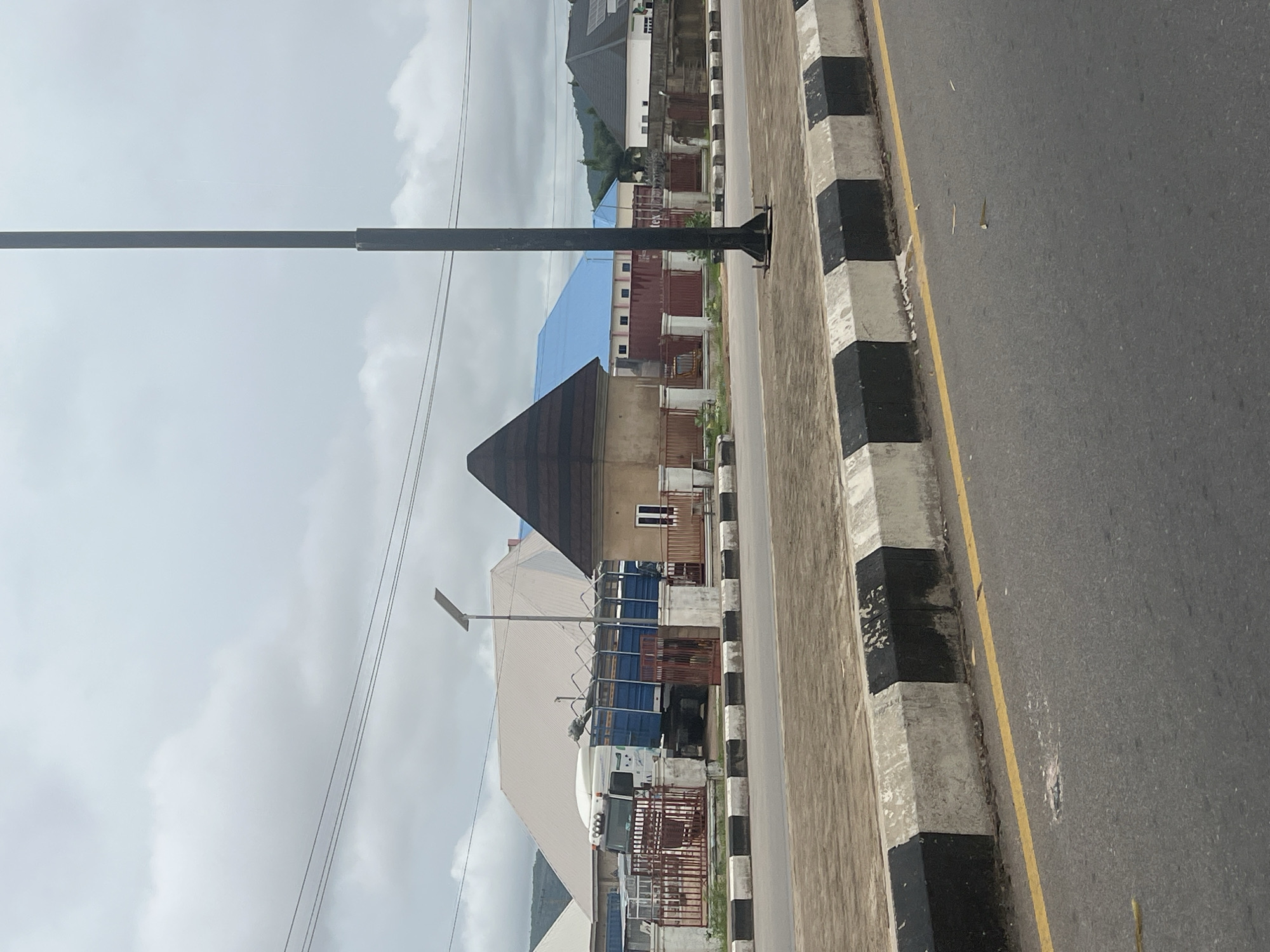 The image depicts a roadside scene in Jalingo, Nigeria. In the foreground, a paved road with a yellow line runs diagonally across the bottom of the frame. A concrete curb with alternating black and white painted blocks separates the road from a sidewalk or paved area. Behind the curb, a large truck with a white cab and a blue cargo bed is parked. The truck has a metal cage-like structure over its cargo bed. The truck is behind a red metal fence.
In the background, several buildings are visible. One building has a distinctive conical, dark-colored roof. To its right, a building with a bright blue roof is seen, with shipping containers behind it. Further to the right, a white building with a dark-tiled roof has solar panels on it. A tall, dark lamppost stands prominently in the right-center of the image. Overhead, power lines are strung across the sky. The sky is overcast with clouds. The overall environment appears to be an urban or semi-urban area, likely along a main road.