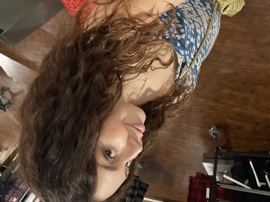 A woman with curly brown hair faces the camera. She wears a blue patterned top with a gold chain strap and matching leaf-shaped bag. A delicate gold necklace adorns her neck. The background features a wooden floor and clothing racks holding red and blue items. The image appears to be taken indoors with warm lighting.