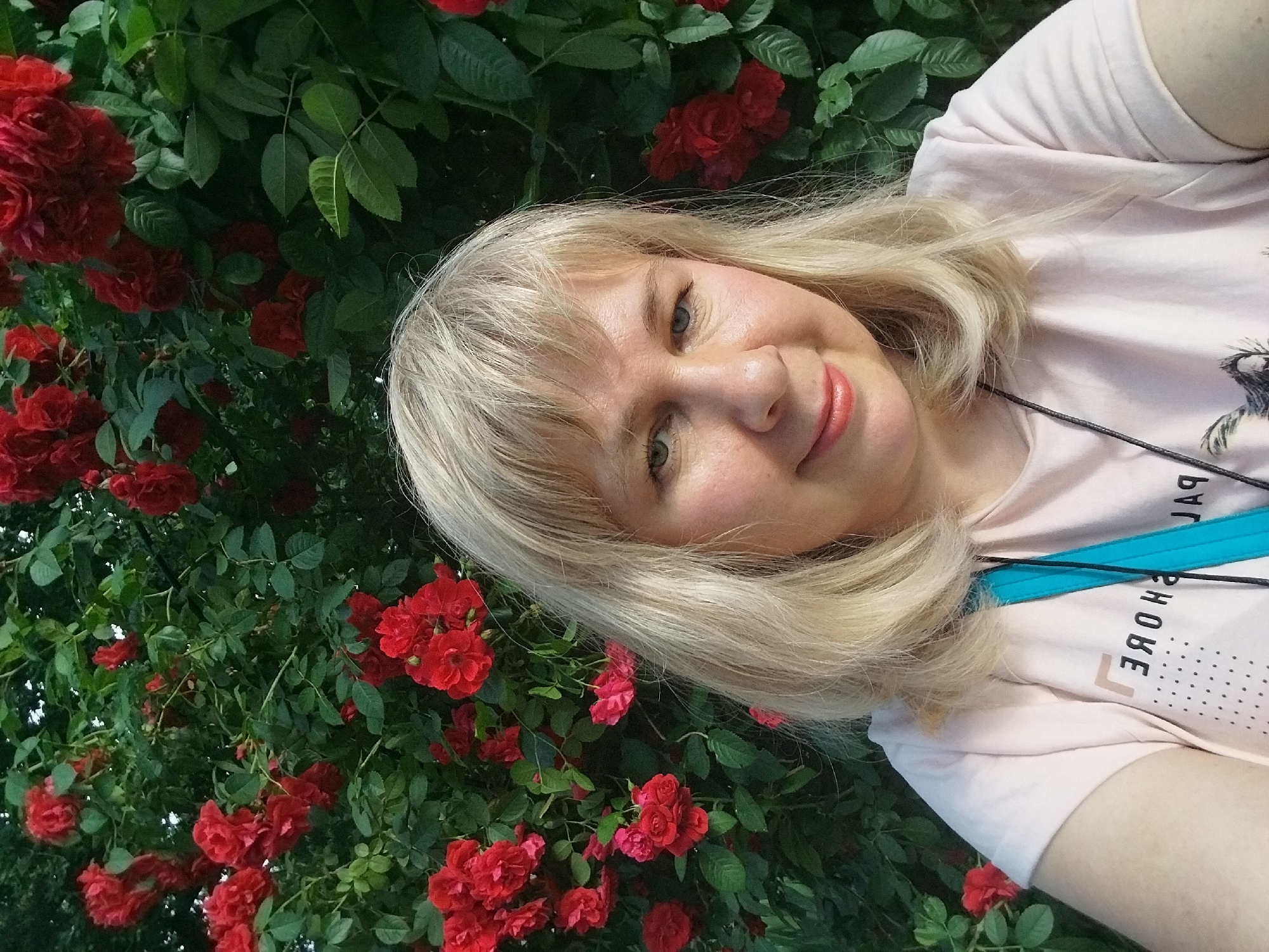 A young woman with blonde hair and blue eyes smiles at the camera. She is wearing a light pink t-shirt with a graphic print on the front. The woman is positioned in front of a bush with bright red roses. The image appears to be taken outdoors, possibly in a garden or park, with the natural light suggesting it was taken during the day. The location is identified as Chernorech'e, Russia, indicating a potentially rural or suburban setting.