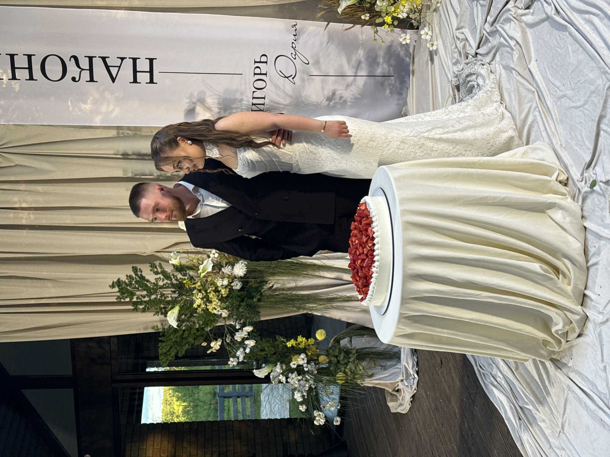 The image captures a wedding couple posing beside a decorated cake and floral arrangements within an event venue. The male, on the left, has a beard and short dark hair, wearing a black suit jacket and a white collared shirt. He is looking towards the left, with his right arm around the female's waist. The female, on the right, has long dark brown hair styled partly up, wearing pearl earrings and a white strapless lace wedding gown with a high lace choker. She is also looking towards the left.
In the foreground, slightly left of center, a white cake generously topped with strawberries rests on a black cake stand, placed on a round table draped in light cream fabric. Behind the couple, a tall white banner displays Cyrillic text: "НАКОН" vertically, and "ИГОРЬ" and "Дария" (Igor and Daria) horizontally in black lettering, signifying the couple's names. The banner also features subtle grey patterns.
Elaborate floral arrangements containing white, yellow, and green flowers, including calla lilies and trailing greenery, are positioned on draped pedestals to the left of the couple and behind the cake table. The background features light-colored curtains or drapes. To the far left, a brick wall and dark wooden architectural elements are visible. Through a large glass doorway or window on the left, an outdoor scene is visible, depicting green trees and a distant fence under what appears to be a bright sky. The flooring consists of wooden planks, partially covered with loosely draped white fabric.
The Cyrillic script on the banner indicates a Russian-speaking region. However, no specific landmarks or identifying features are present in the image to forensically pinpoint the exact location within the city of Perm, Russia.