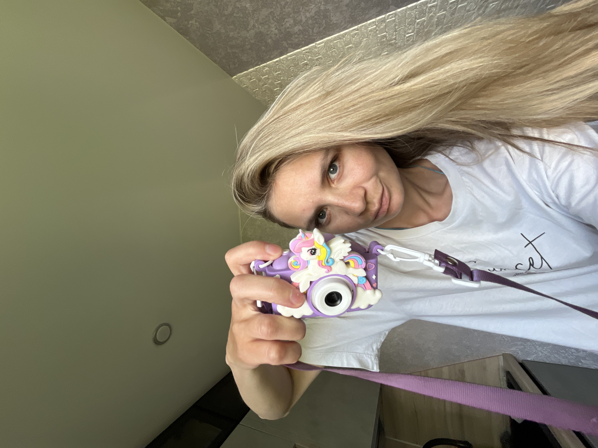 A blonde woman in a white t-shirt is holding a purple and white toy camera with a unicorn design in front of her. She appears to be taking a selfie. The background shows a plain light-colored wall, a light fixture on the ceiling, and a patterned section of ceiling. The location is Chaikovsky, Russia.