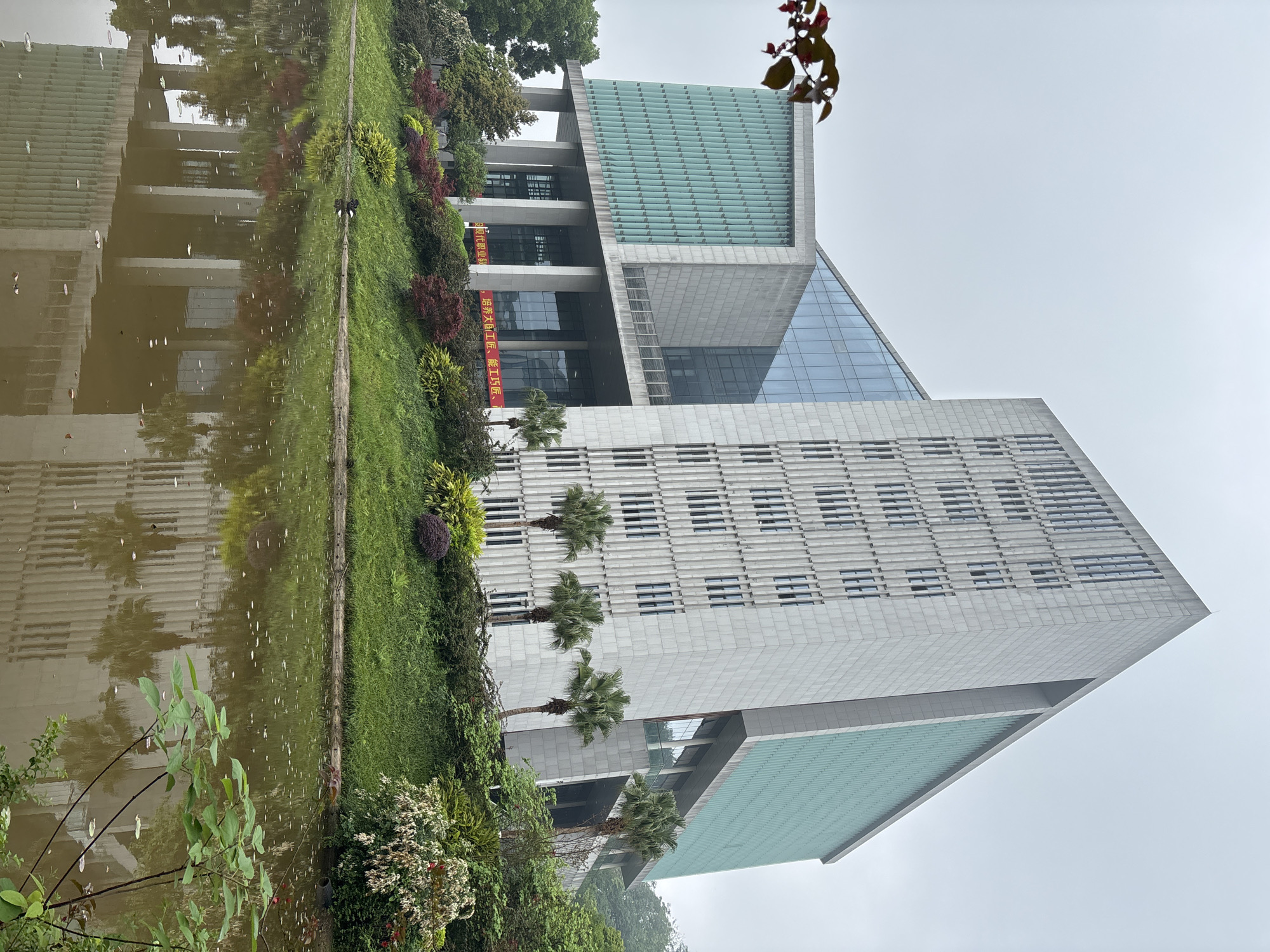 The image shows a modern building next to a body of water, possibly a river or a pond. The building has a gray facade and many windows, with a green roof section. There are palm trees and greenery along the bank between the building and the water. The water reflects the building and the surrounding vegetation. The sky is overcast. The building's architecture is angular and appears to be a modern or contemporary design. Banners with Chinese characters are affixed to the side of the building. The photograph was taken in Jiangjin, China.