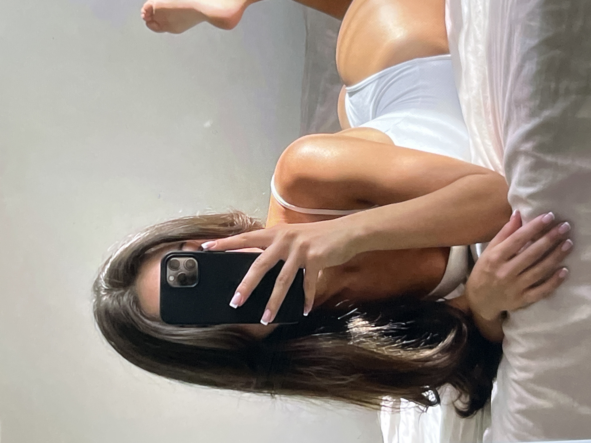 A young woman with long, dark hair lies on a bed, taking a selfie with her phone. She is wearing a white one-piece swimsuit. Her right arm is extended, holding the phone up to take the picture, while her left arm is bent at the elbow and rests on the bed. Her face is obscured by the phone. The background is a plain, light-colored wall. The lighting suggests it is daytime, with natural light coming from the side, creating highlights on her skin. The overall setting appears to be a bedroom.