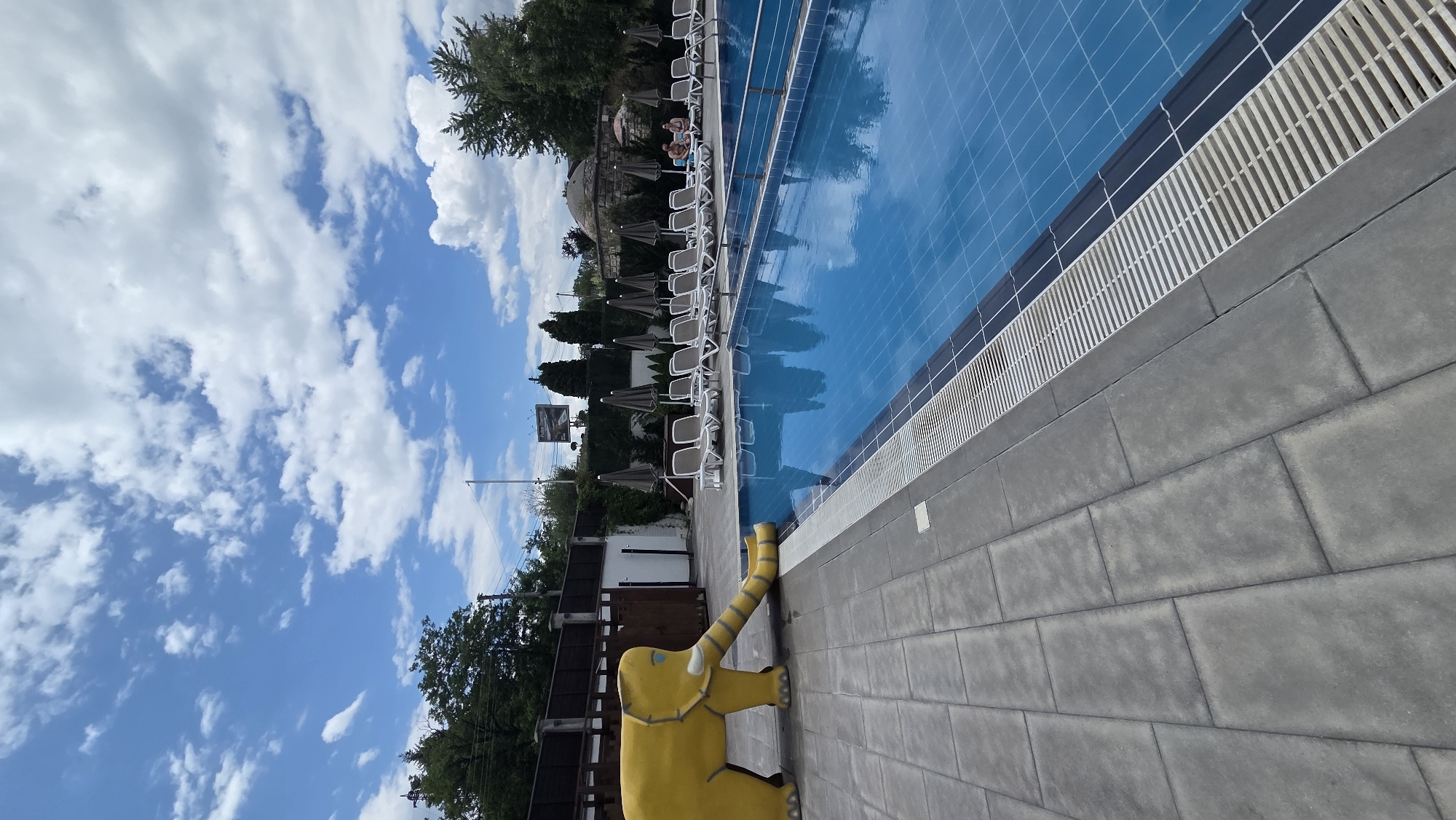 This media file shows an outdoor swimming pool area in Bania, Bulgaria. The pool is bright blue and reflects the sky, which is partly cloudy with white cumulus clouds against a clear blue backdrop. Sunlight suggests it is daytime, likely a warm, pleasant day.
The pool is lined with white lounge chairs and umbrellas, some of which are open, suggesting people are relaxing or have recently been present. A few individuals can be seen sitting in lounge chairs near the pool. Adjacent to the pool is a paved area made of large, grey rectangular stones. In the foreground, a bright yellow, cartoonish elephant statue is positioned on the paved area, with its trunk extended upwards.
Further back, behind the lounge chairs, is a line of trees and some fencing. There is a sign visible in the distance, though the text is not legible. The overall setting appears to be a recreational facility, possibly a resort or a public pool, designed for relaxation and leisure. The presence of lounge chairs, umbrellas, and the swimming pool indicates an environment suited for enjoying sunny weather.