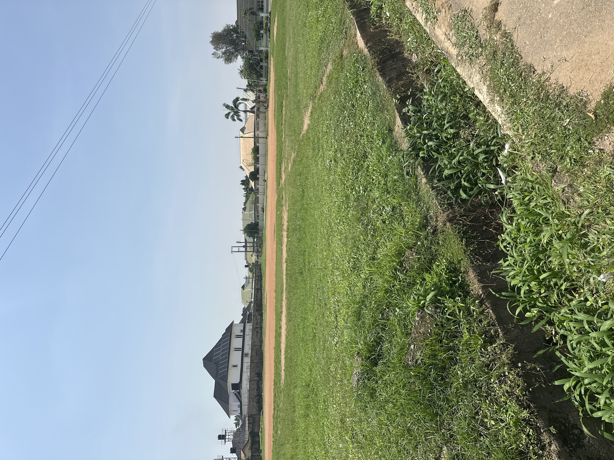 The image depicts an open, grassy field in Abuja, Nigeria, under a clear, bright sky. In the background, a white house with a dark, multi-pitched roof, likely covered in solar panels, stands prominently behind a concrete fence. Several other residential buildings with cream and beige roofs are visible further in the distance, alongside trees and utility poles. A dirt track or path runs across the middle ground of the field, separating two grassy areas. In the foreground, a concrete drainage ditch filled with lush green vegetation runs along the right edge of the image, bordering a paved walkway or road. The overall scene suggests a suburban or semi-rural residential area.