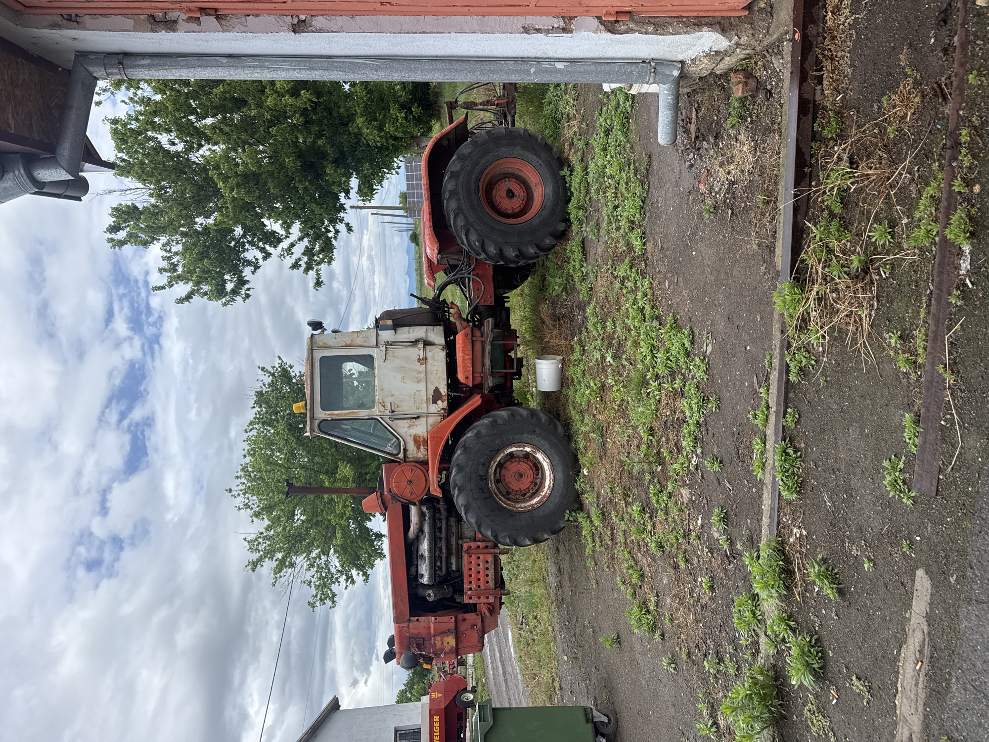 The image shows a dilapidated, likely non-operational, orange and white tractor parked on a paved area with dirt and vegetation. The location appears to be outdoors, possibly near a building with a red brick or tile roof. A tree is visible in the background, partially obscuring the sky, which is overcast. To the right of the tractor, a metal drainpipe is attached to the building. In the foreground, part of a green container and what appears to be a red farm implement are visible. There is no visible activity or people. The location might be in or near Sarnevo, Bulgaria based on information included.