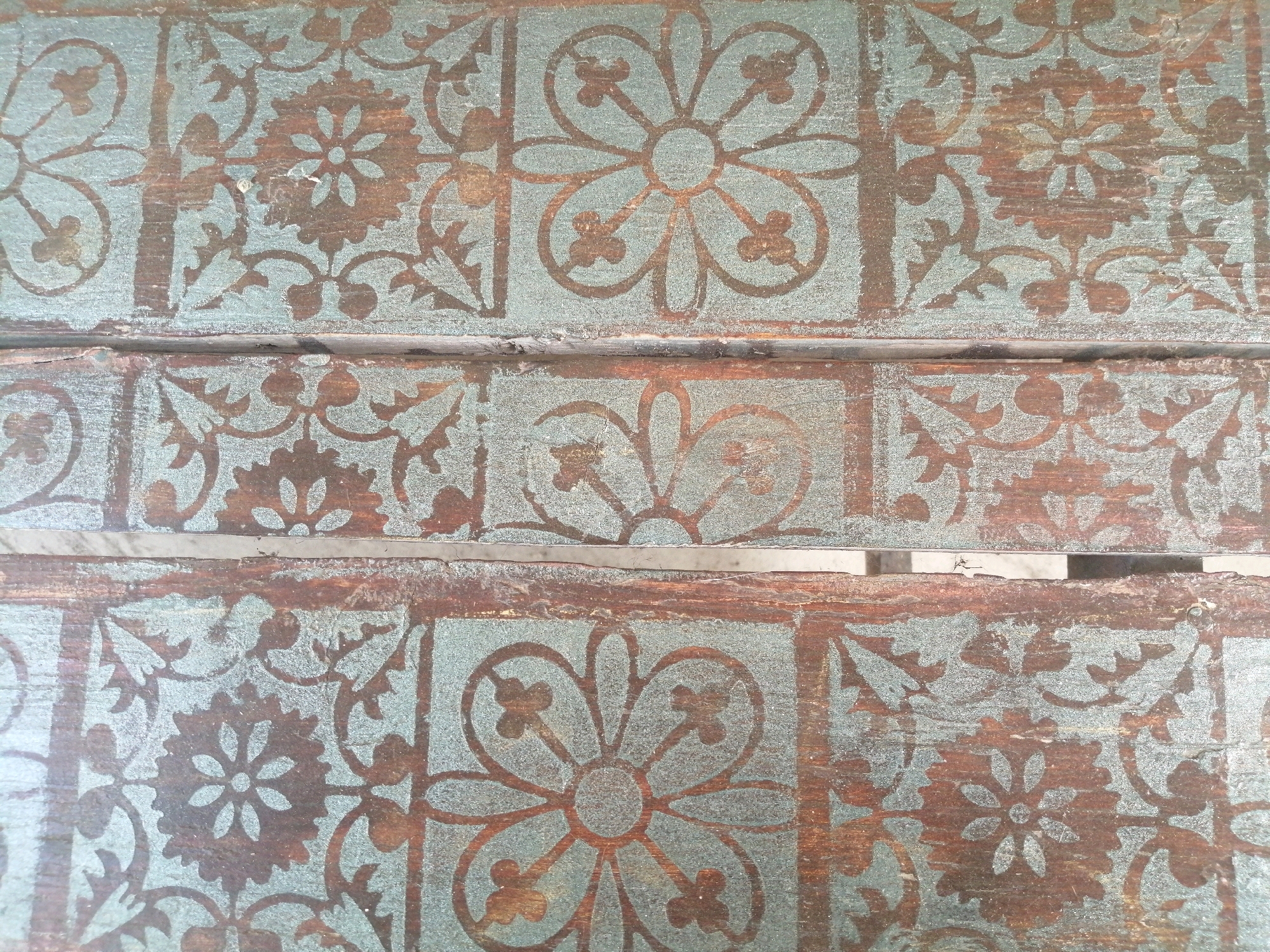 The image shows a close-up, overhead view of a weathered wooden surface with a repeating pattern stenciled onto it. The pattern consists of floral motifs and geometric elements in a dark brown or reddish-brown color, overlaid on a distressed teal-colored paint. The wood itself appears to be planks, with visible grain and signs of age, such as scratches and chipping paint. The pattern is divided into square or rectangular panels by a horizontal line running across the middle of the image, suggesting it might be a tabletop, a bench, or a decorative panel. The lighting is even, and there are no people or other discernible objects in the scene. The location is Unknown, Unknown.