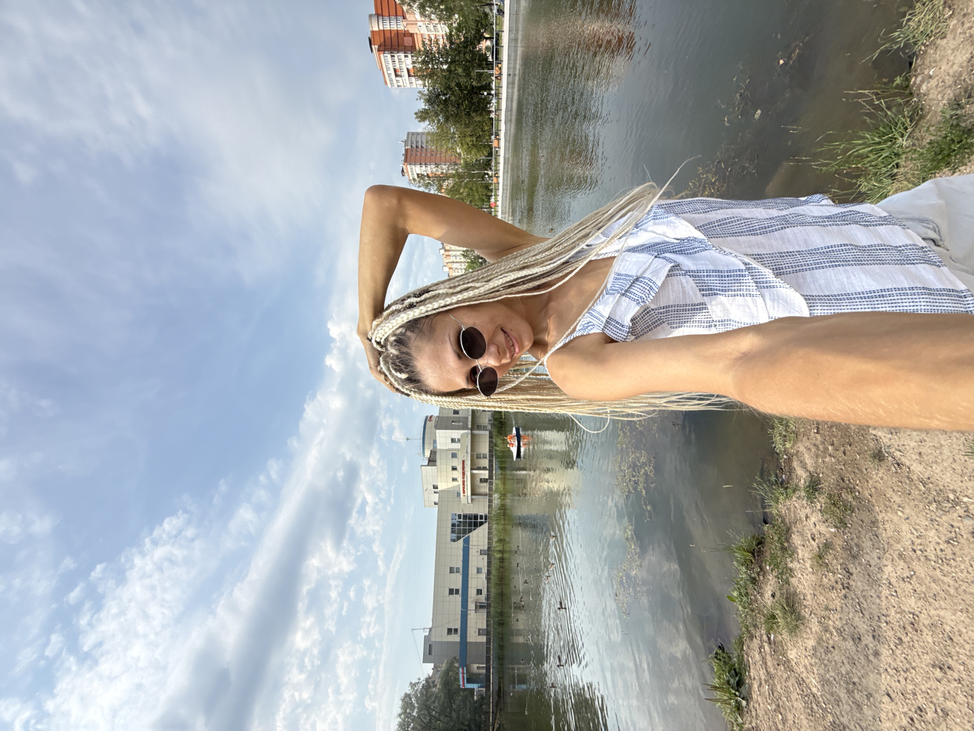 A woman with blonde braids and sunglasses is taking a selfie by a lake with buildings in the background. She is wearing a white and blue striped top and white shorts. The lake is calm and reflects the sky and buildings. There are ducks in the water and a small paddle boat with two people in it. The environment is an urban park with trees and grass. The sky is blue with some clouds. The buildings in the background appear to be residential and commercial. The image is taken during the daytime.