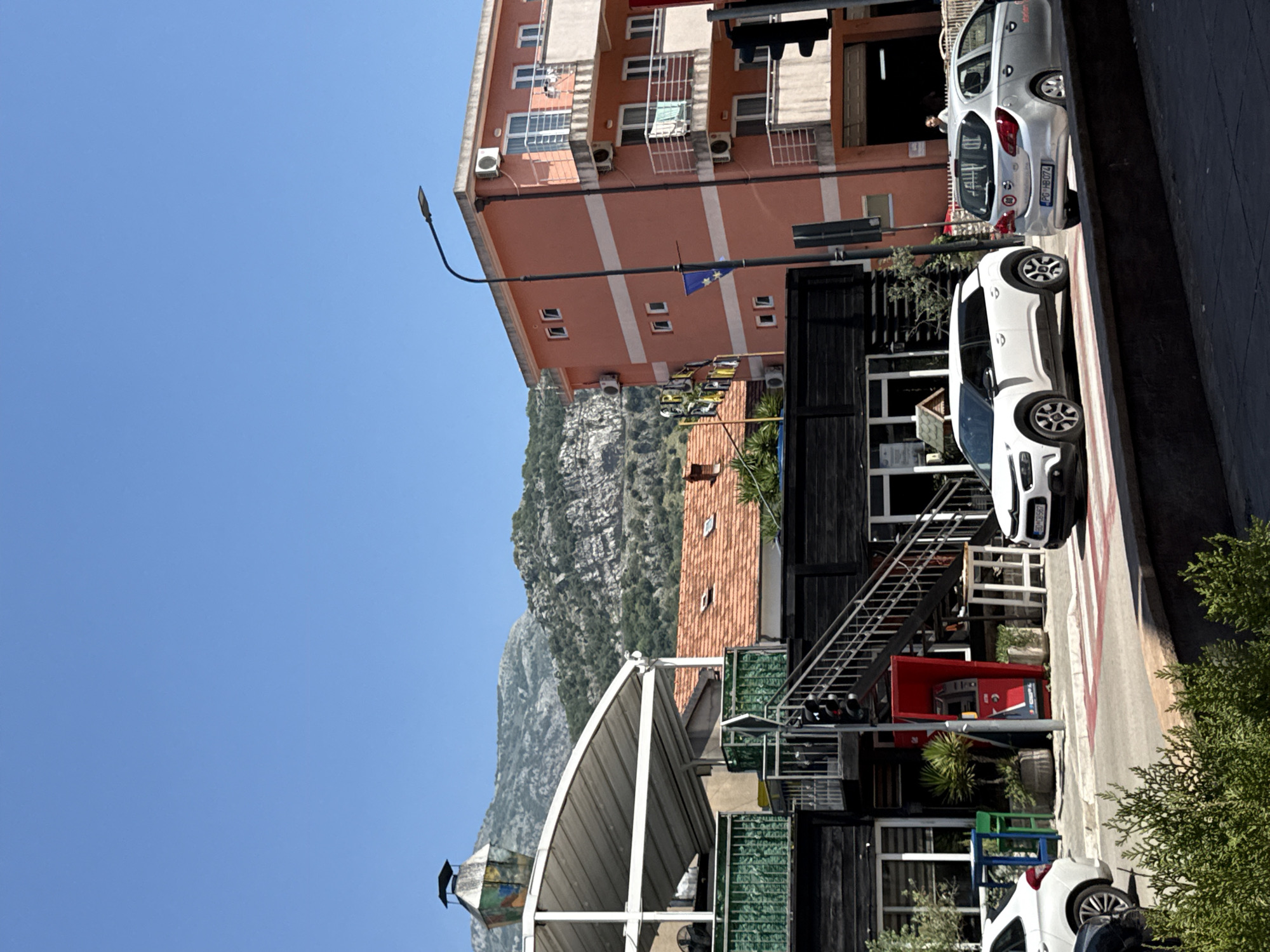 This is a street scene in Budva, Montenegro, on a clear, sunny day. The foreground features a pedestrian crossing with red and white stripes. To the right, a silver hatchback car with license plate "PG HB074" is parked on the street. To the left, a white crossover SUV is also parked on the street, with a portion of its rear bumper visible.
Behind the cars, a dark-colored, two-story building with large windows is visible. An outdoor staircase with metal railings leads up to the second floor. Next to the staircase, a red ATM is housed within a protective structure. In front of the building, there are several tables and chairs, some of which are colored blue and green, and potted plants.
Further back, a light-colored structure with a large, angled canopy is partially visible, topped with a decorative, lantern-like object. Adjacent to this is a building with an orange-tiled roof.
To the right of these buildings, a salmon-pink, multi-story building stands prominently. It has multiple windows and balconies with white railings. Air conditioning units are visible on its exterior. A street light with a curved arm extends over the scene, and a small flag, possibly the European Union flag, is attached to the pole.
In the background, a range of rocky, tree-covered mountains under a clear blue sky dominates the upper portion of the image. The overall impression is one of a vibrant urban environment with natural scenery.